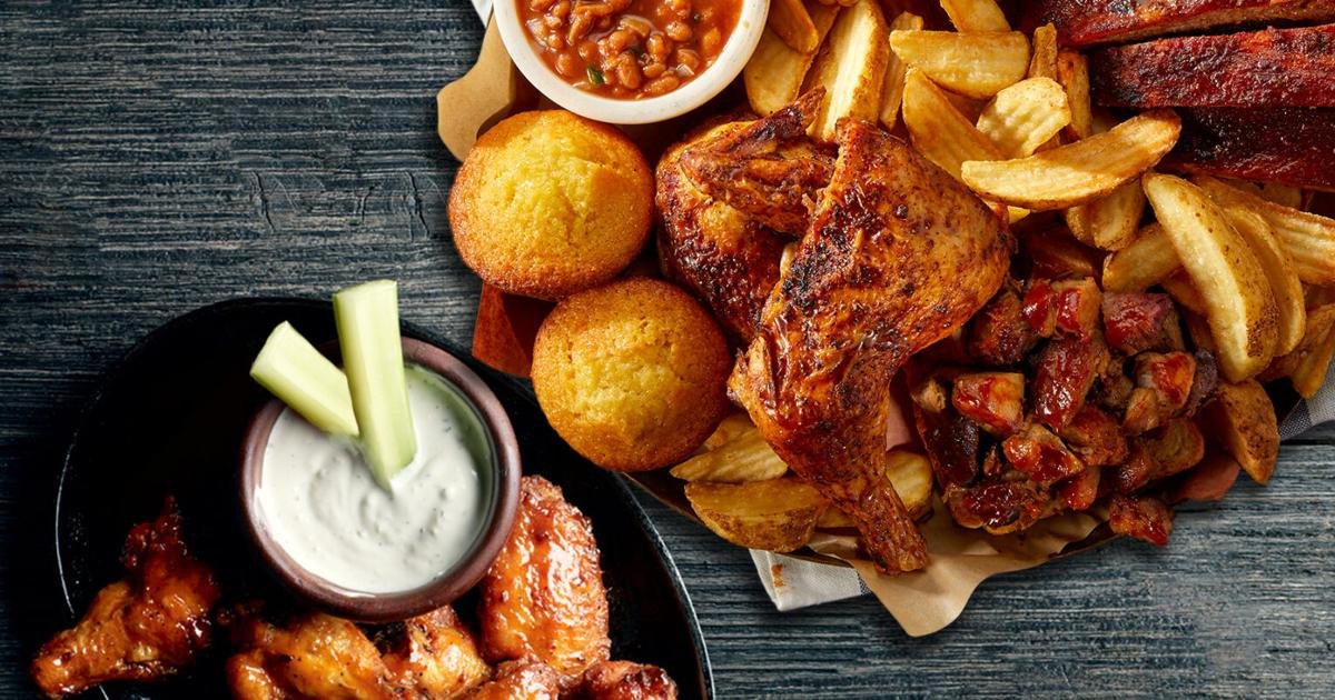 Overhead view of a barbecue meal with ribs, chicken, fries, cornbread muffins, and baked beans on a dark wooden surface.