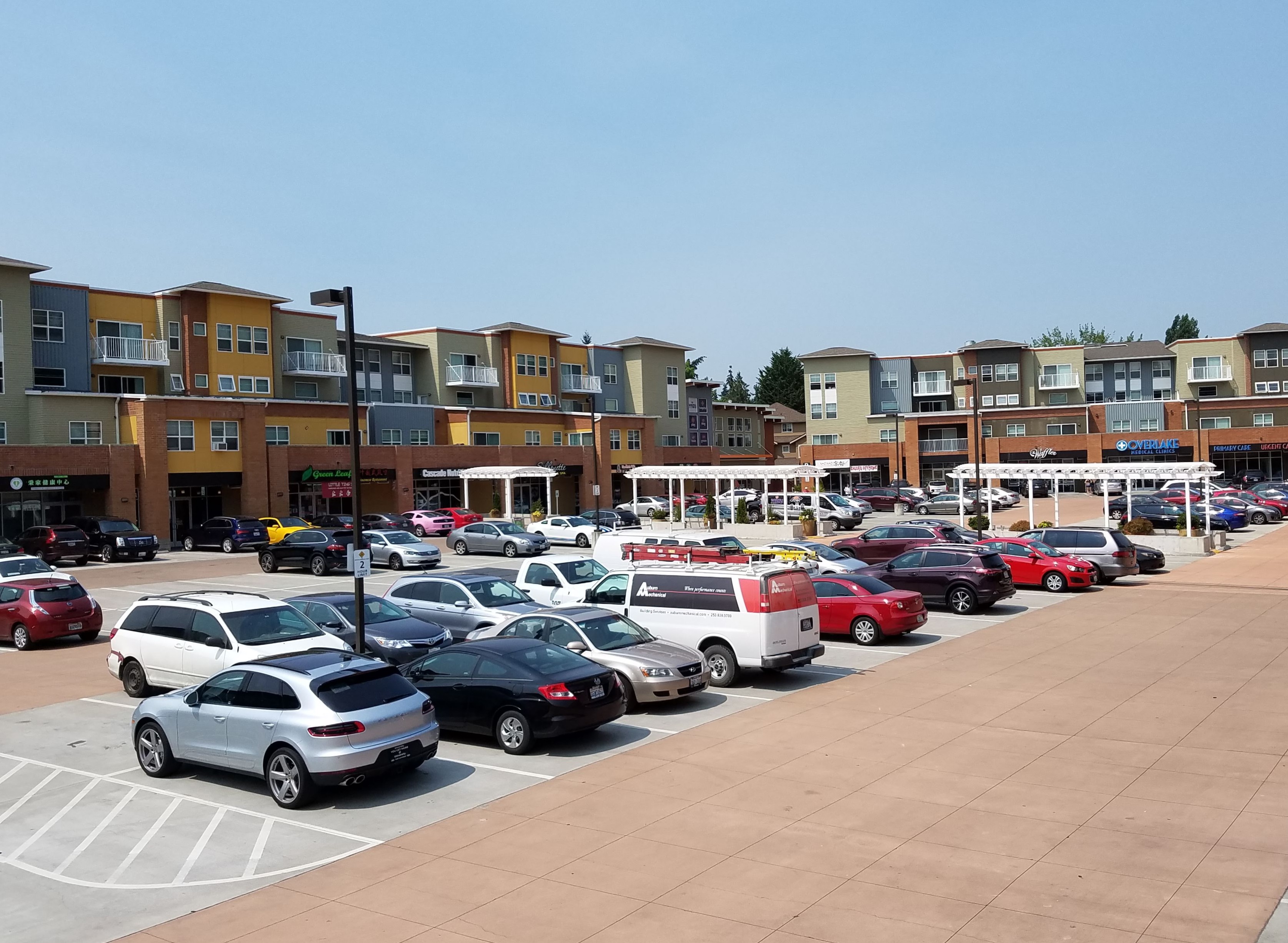 Parking lot in front of a multi-story mixed-use building with shops on the ground floor and residences above.
