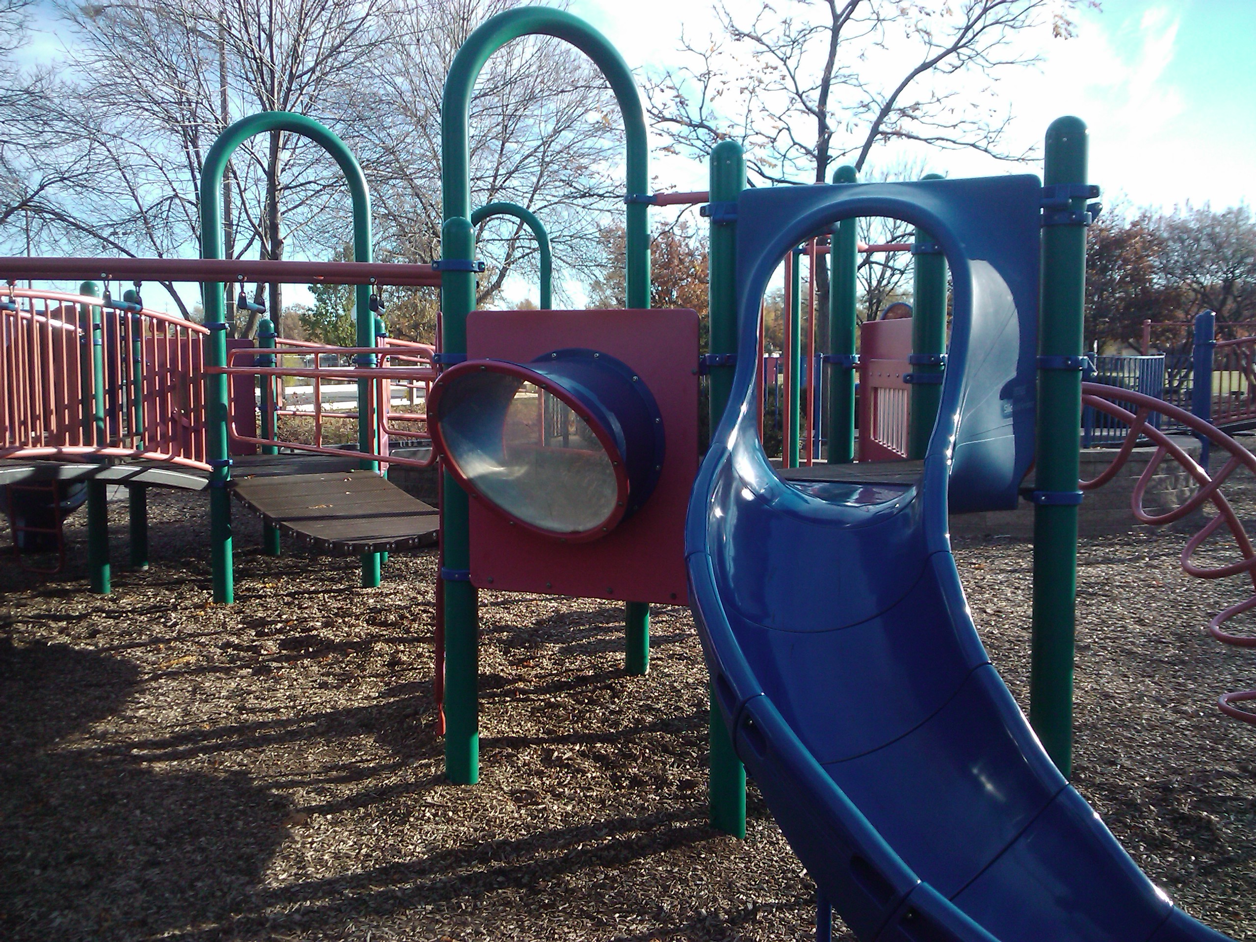 Colorful playground structure with blue slide and green climbing bars on wood chip ground cover.