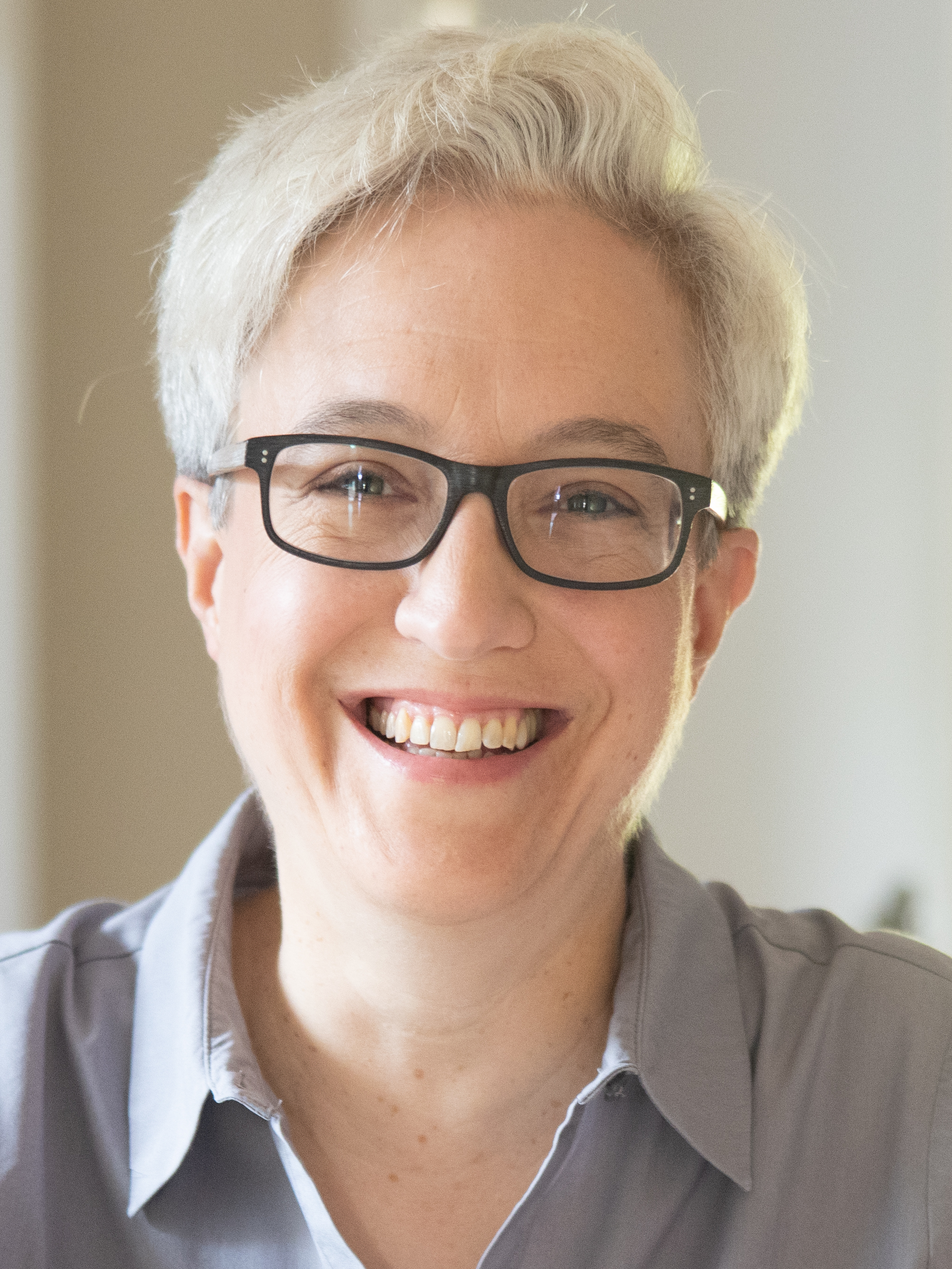 Smiling person with short white hair and dark rectangular glasses wearing a light gray shirt.