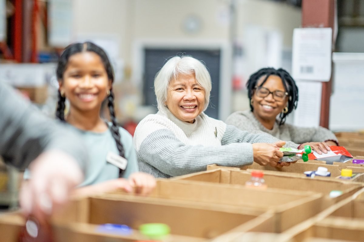 Three smiling women sort colorful items into cardboard boxes at a warehouse table.