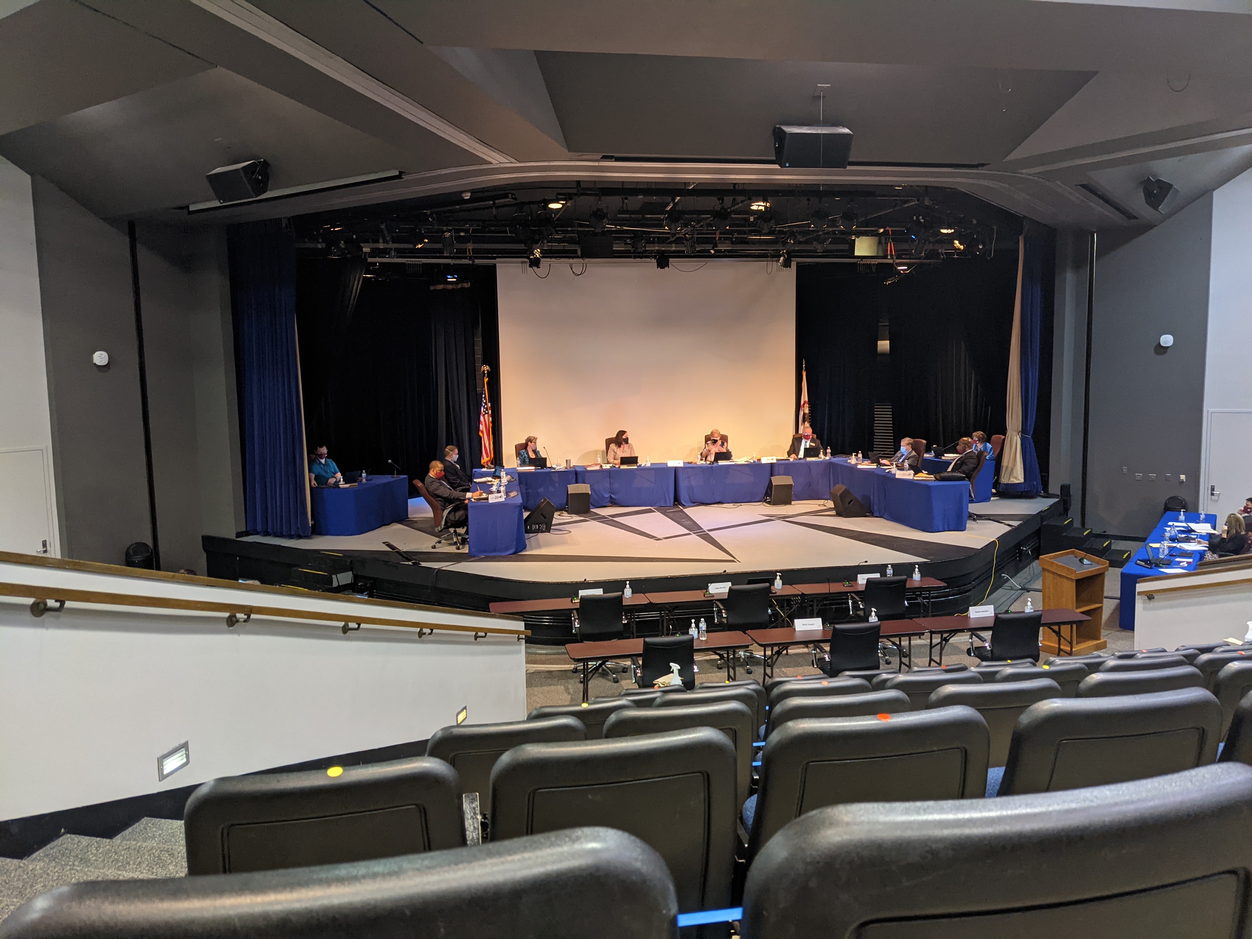 School board members seated at a long table on an auditorium stage during a meeting.