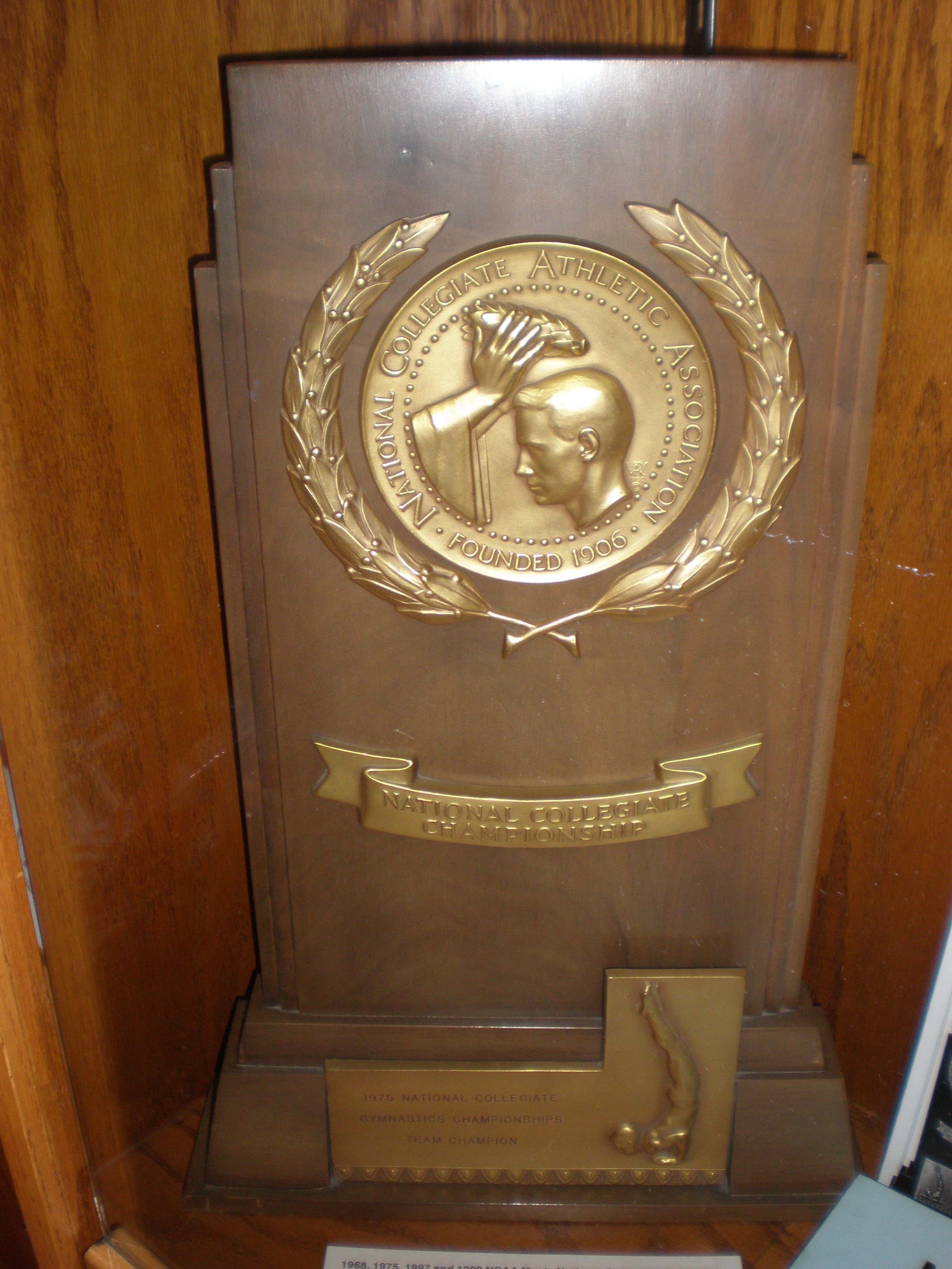Bronze NCAA gymnastics championship trophy mounted on a wooden plaque in a display case.