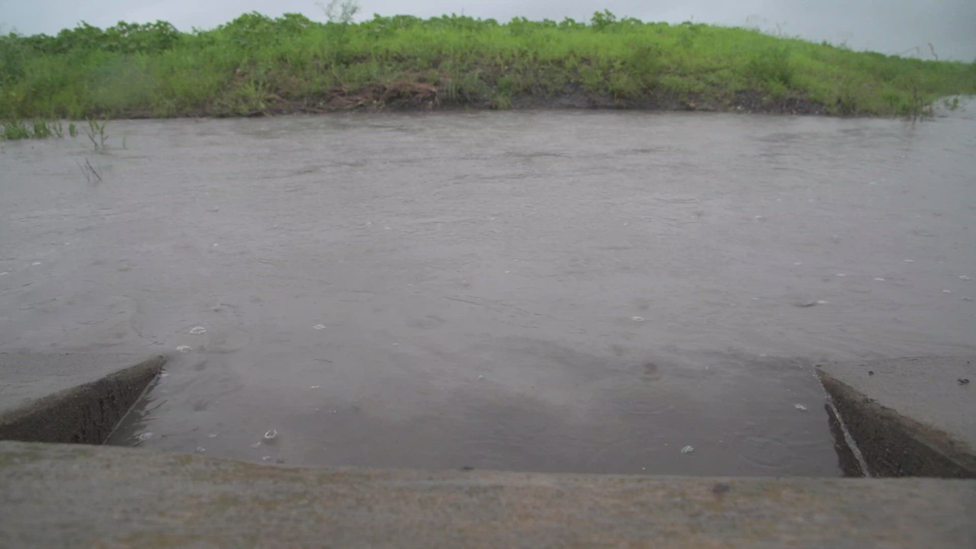 Concrete culvert channeling brown floodwater across a saturated field.