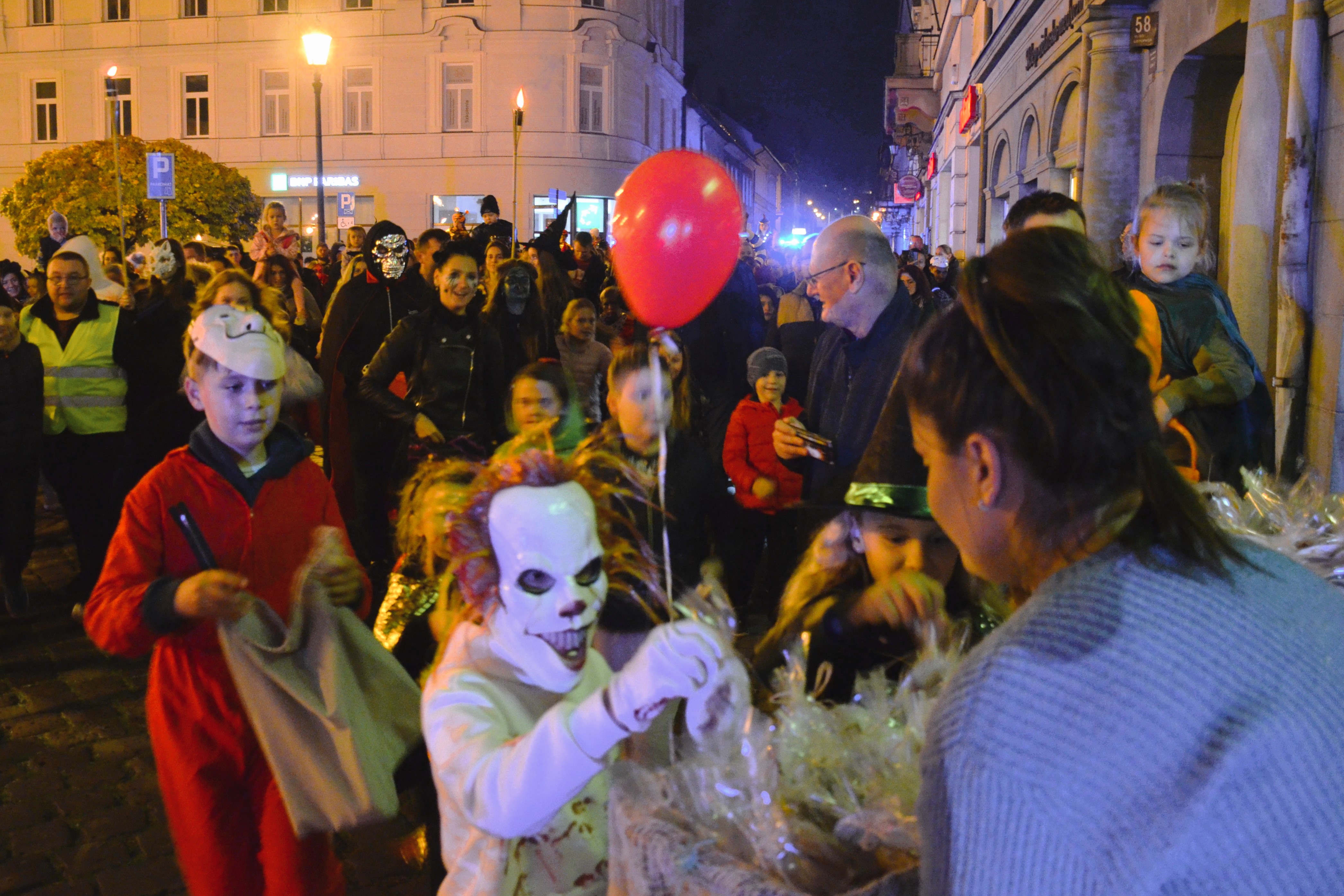 Crowd in Halloween costumes on a city street at night, child in clown costume holds red balloon.