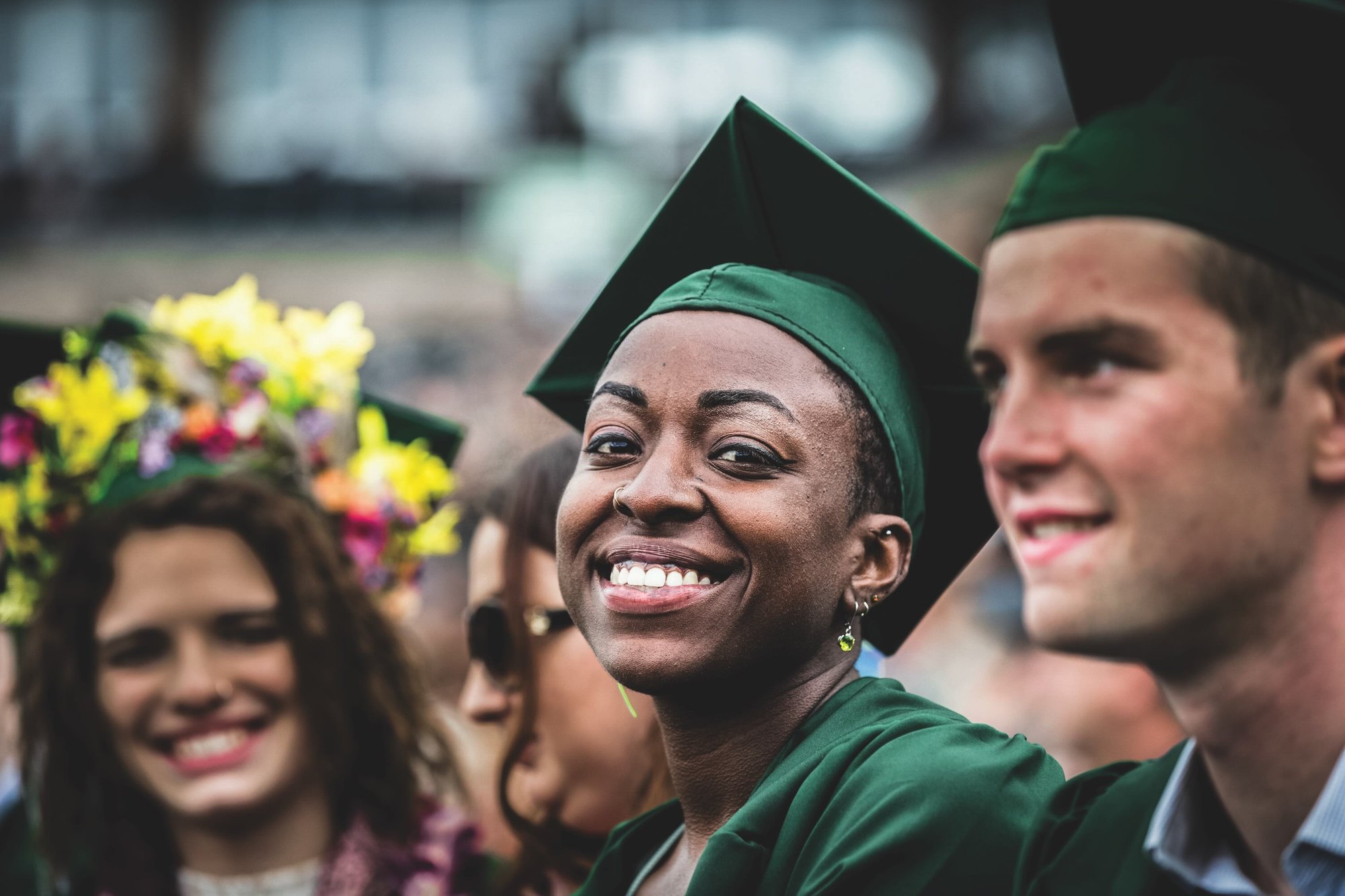 Three smiling graduates wearing green caps and gowns at an outdoor ceremony.