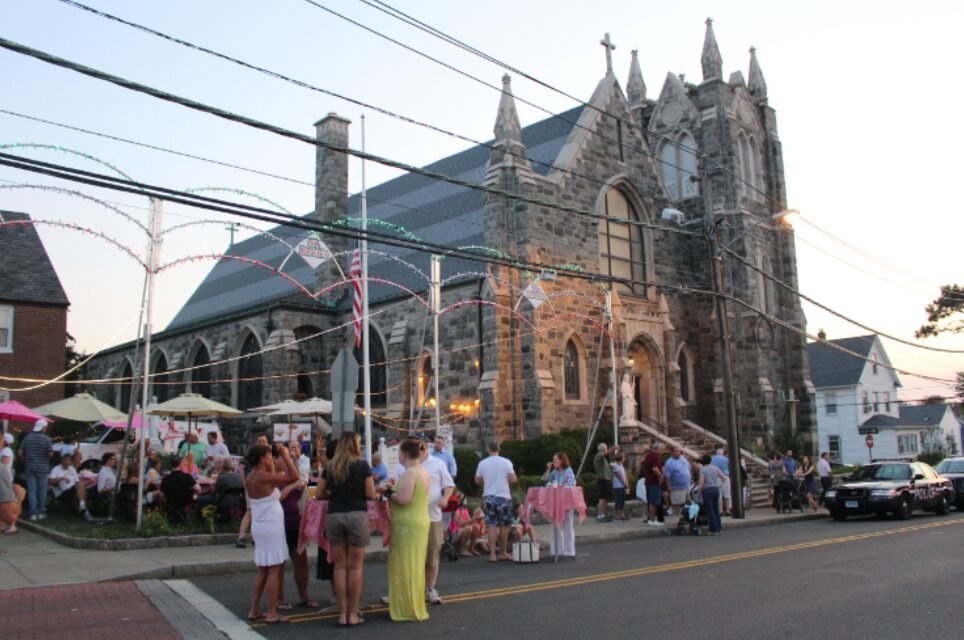 Gothic stone church lit with string lights and bunting, crowd gathered on street at dusk.