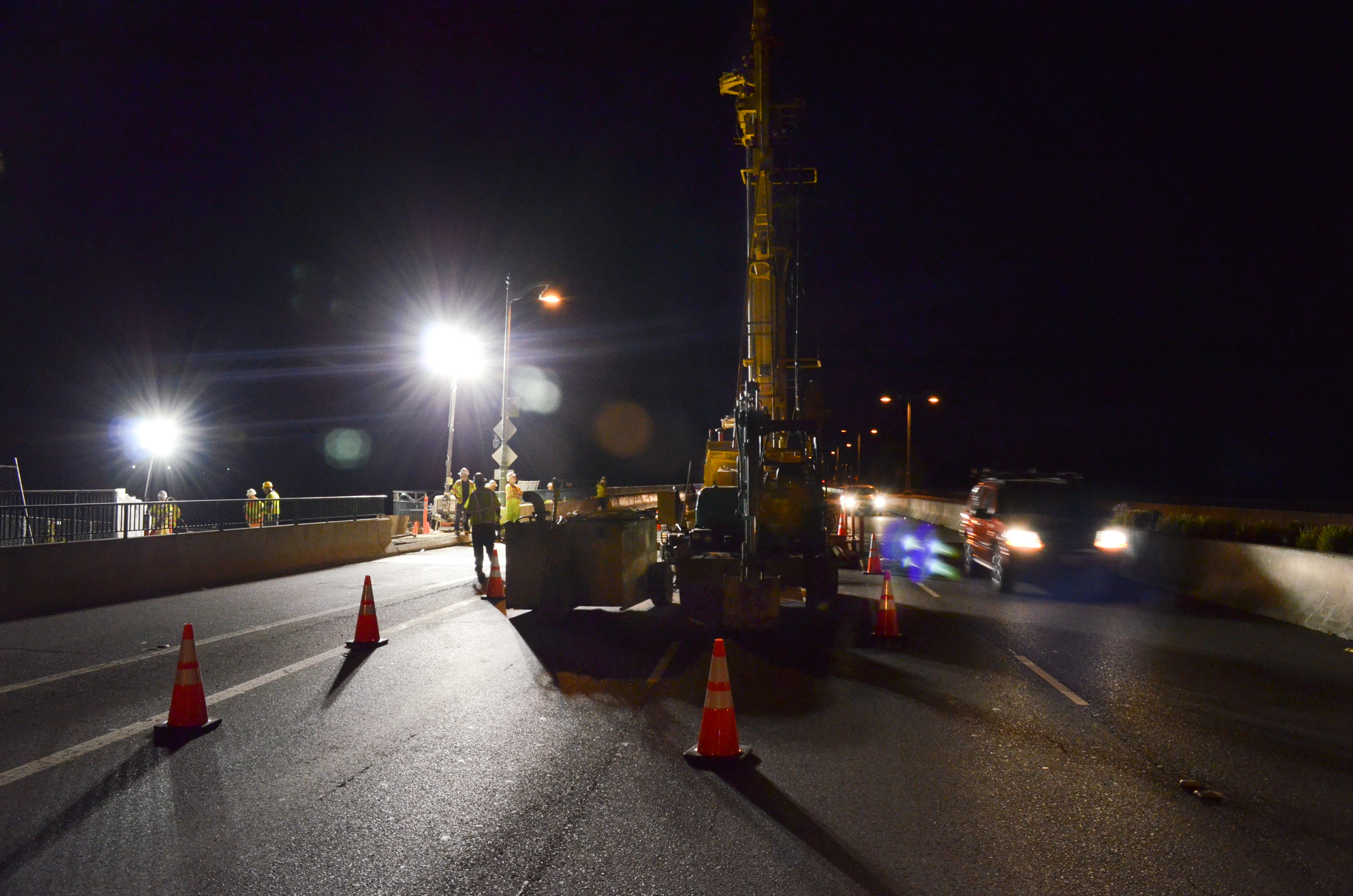 Nighttime road construction with orange cones, a crane, and workers in safety vests on a bridge.