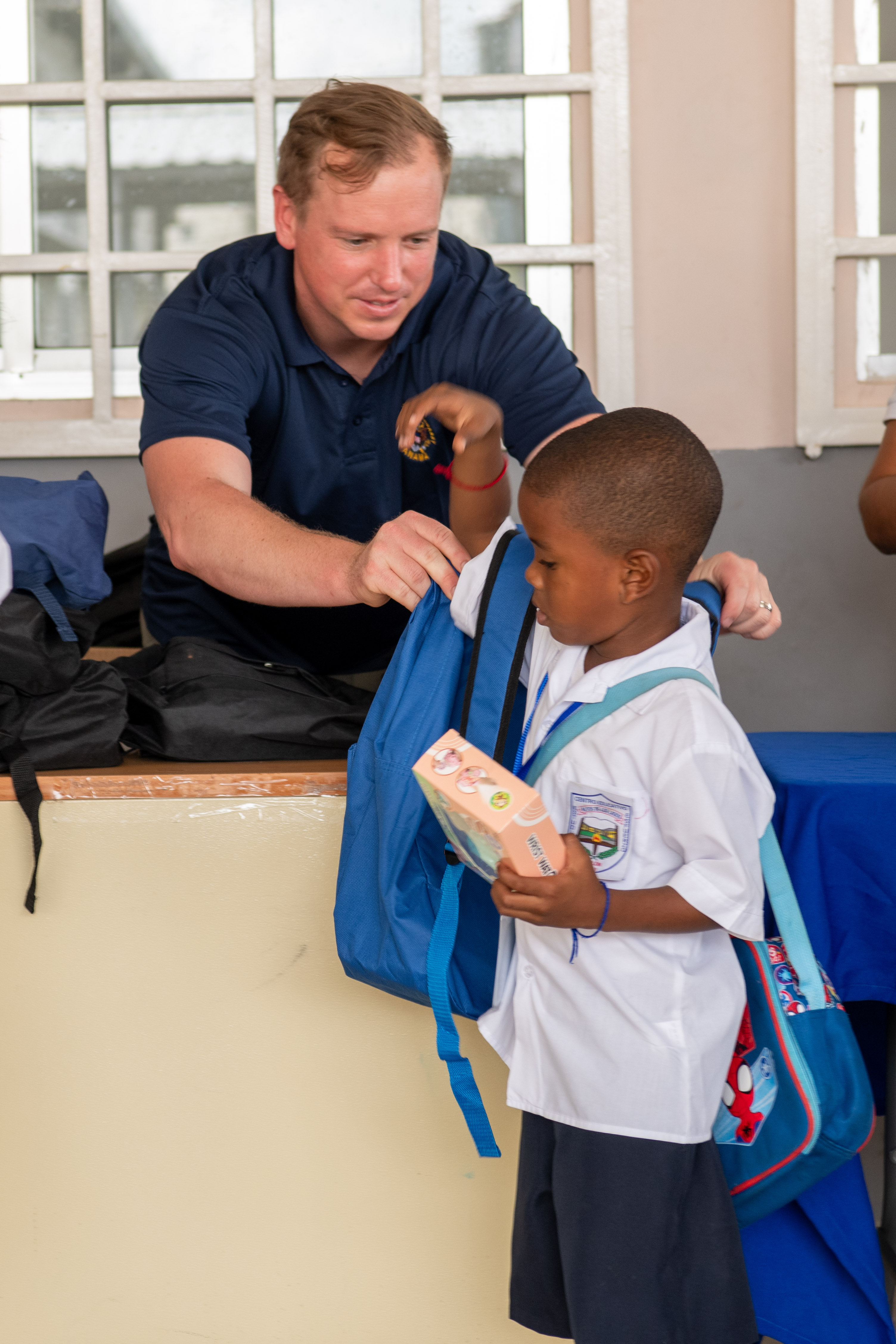 An adult places a blue backpack on a young child inside a school building.