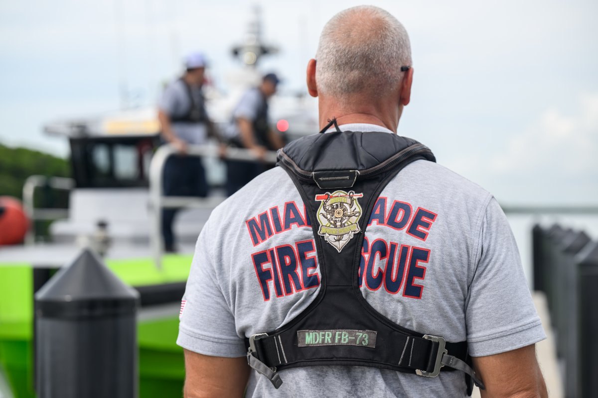 A firefighter in a tactical vest labeled MDFR FB-73 stands near a waterway with a vessel in the background.