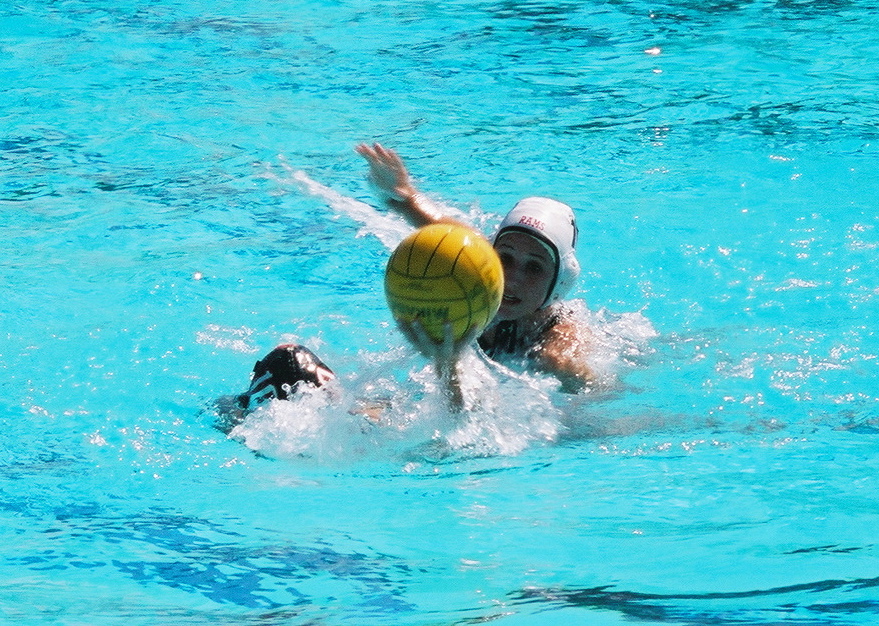 Two water polo players in white caps compete for a yellow ball in a blue pool.