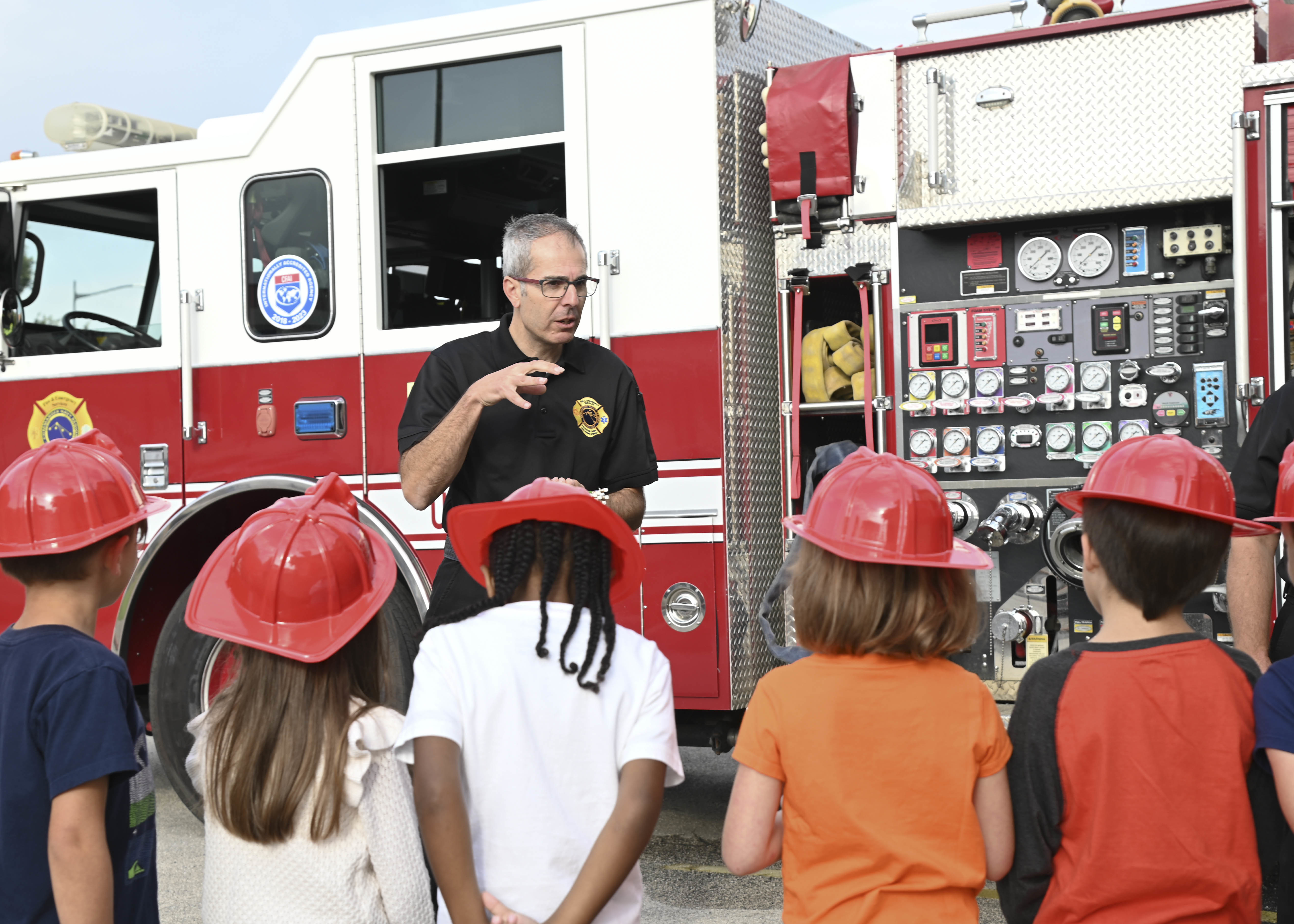 Fire official speaks to children in red plastic helmets beside a red fire truck.