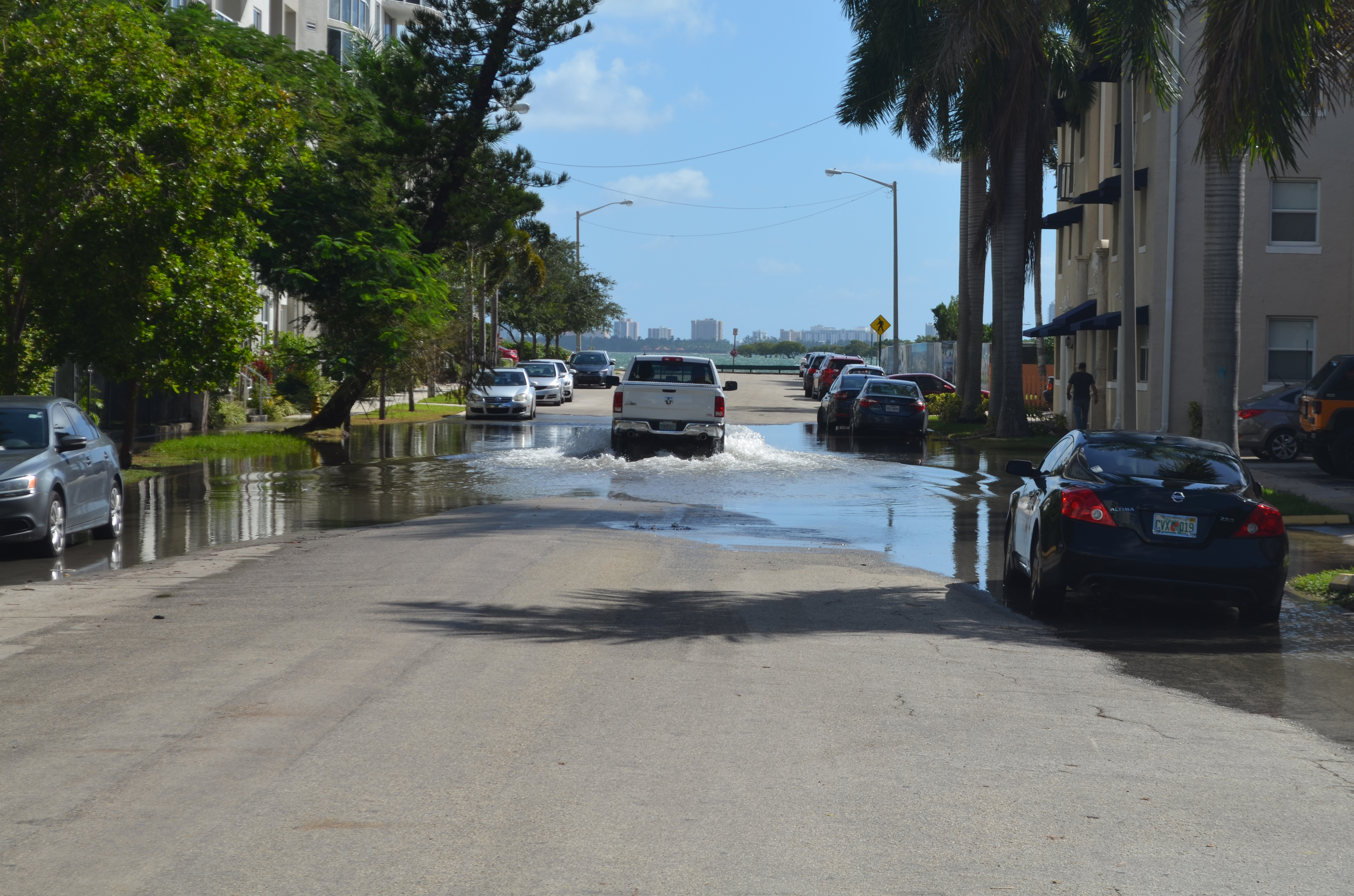 A pickup truck drives through standing water on a flooded urban street lined with palm trees.