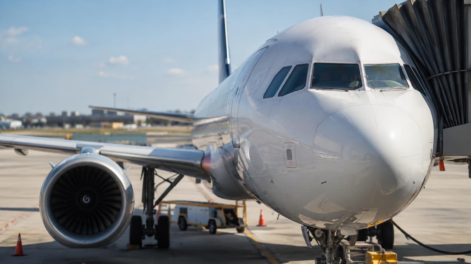 A commercial passenger jet viewed through a terminal gate window on a sunny tarmac.