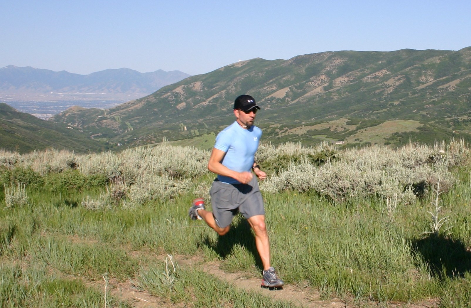 A runner in a light blue shirt and cap runs along a dirt trail through shrubby mountain terrain.