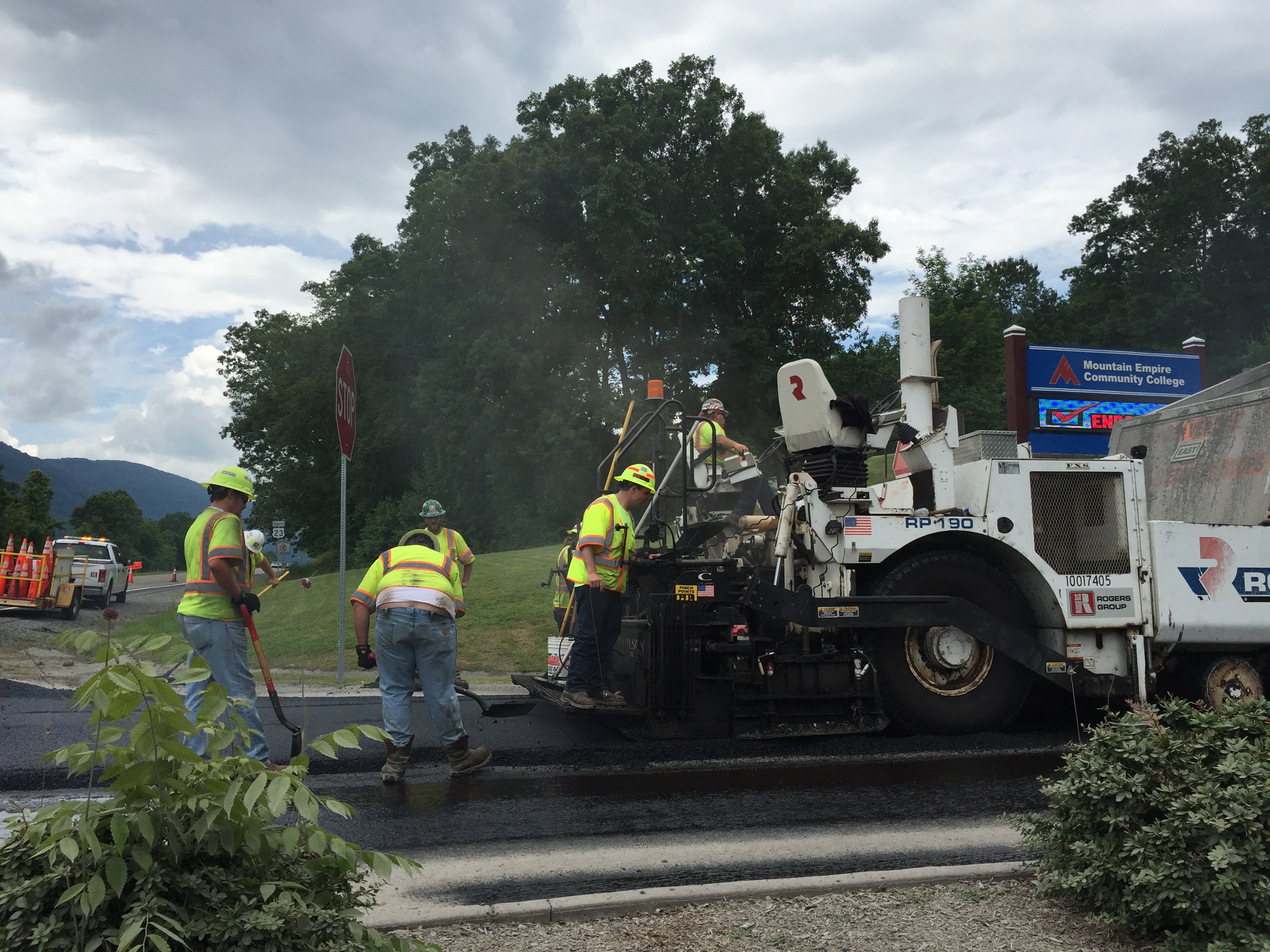 Workers in high-visibility vests operating an asphalt paving machine on a road.