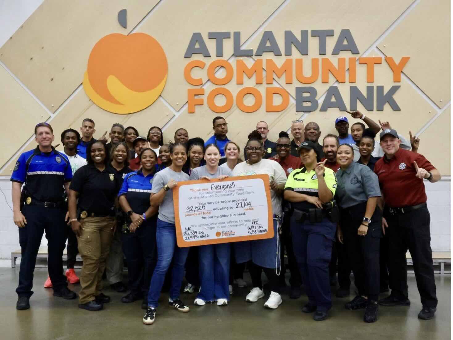 Group of law enforcement officers posing with an oversized check in front of a food bank sign.