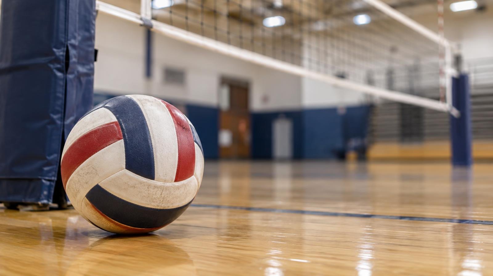 A volleyball resting on a hardwood gymnasium floor near a net post.