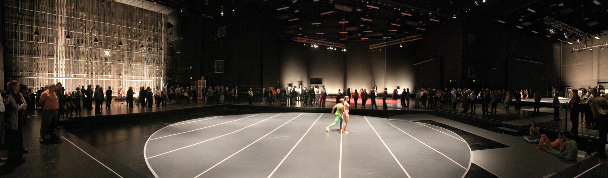 A performer stands on a circular lit stage in a large theater with spectators lining the walls.