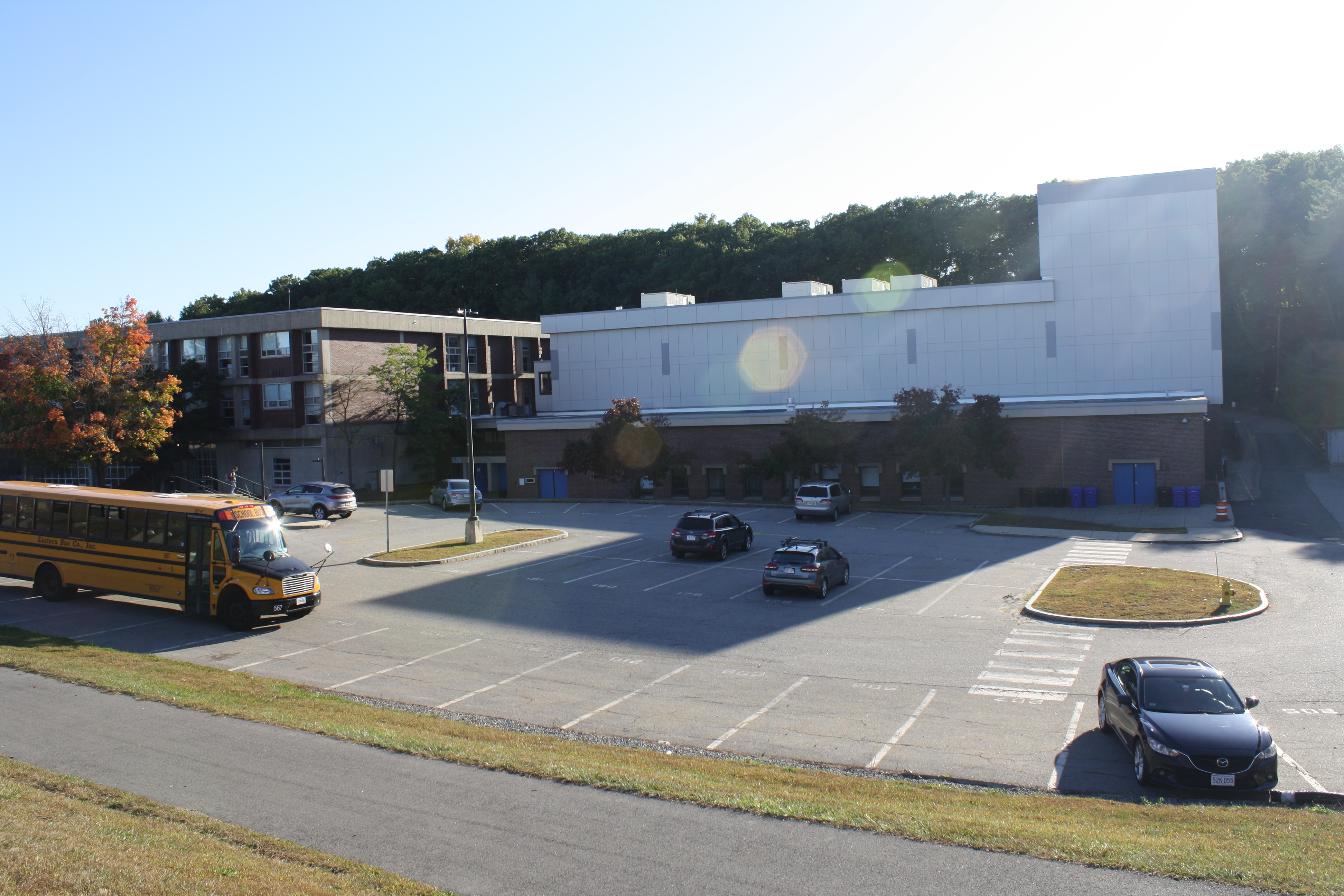 Exterior of a large school building with a parking lot and a yellow school bus, surrounded by autumn trees.