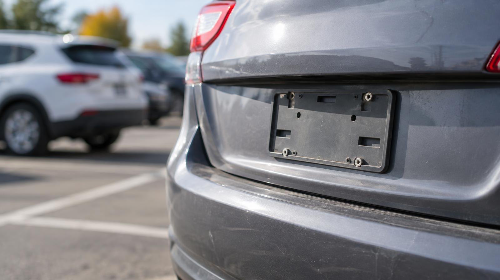 Empty license plate mounting bracket on a car's rear bumper in a parking lot.