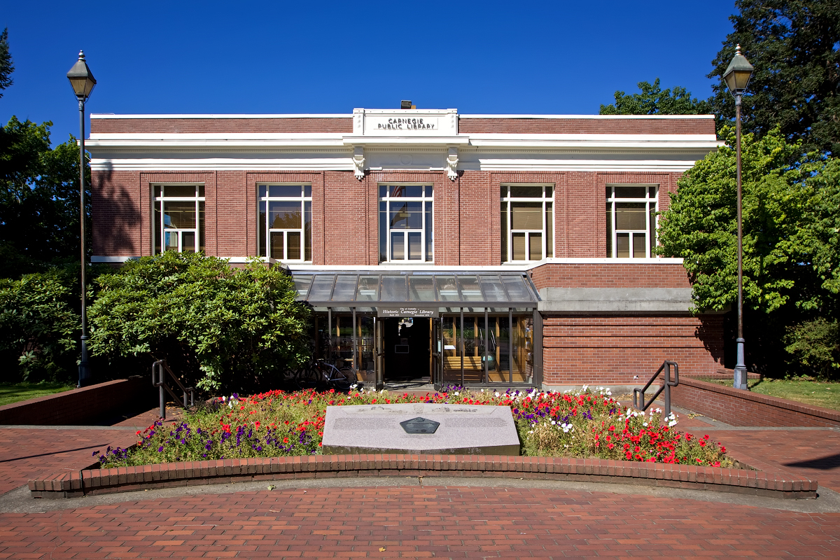 Brick library building with a glass entrance canopy and colorful flower beds.