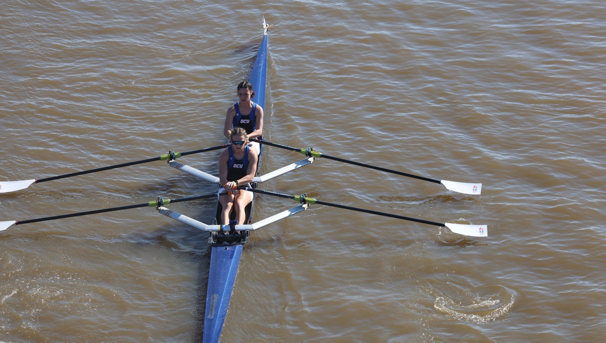 Two rowers in a blue double scull on brown river water, viewed from above.