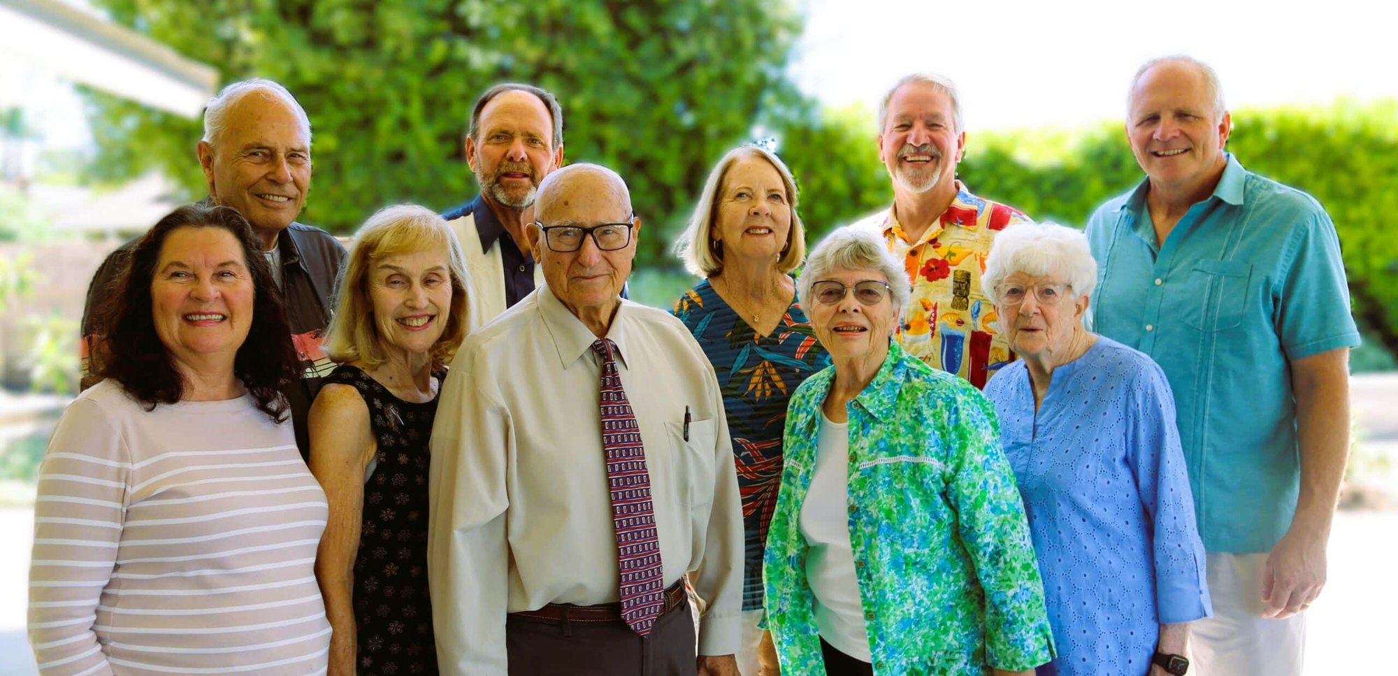 Ten older adults, men and women, posed together outdoors in front of green foliage.