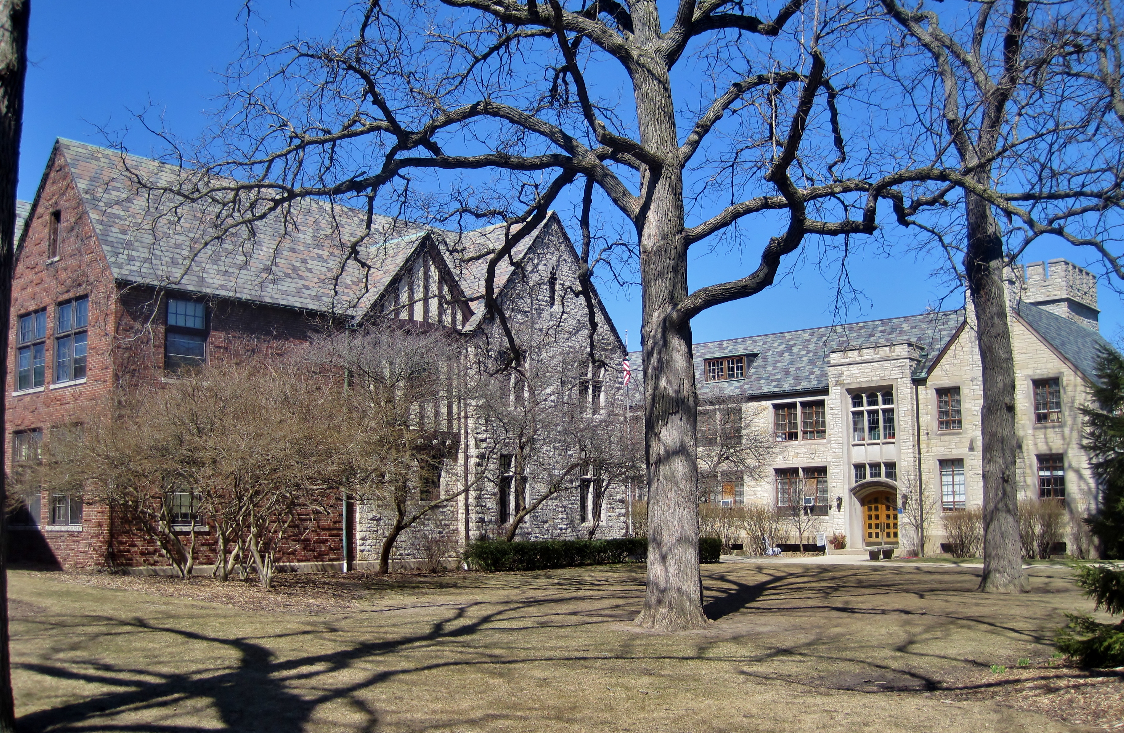 Two Tudor-style stone buildings surrounded by bare winter trees on a grassy lawn.