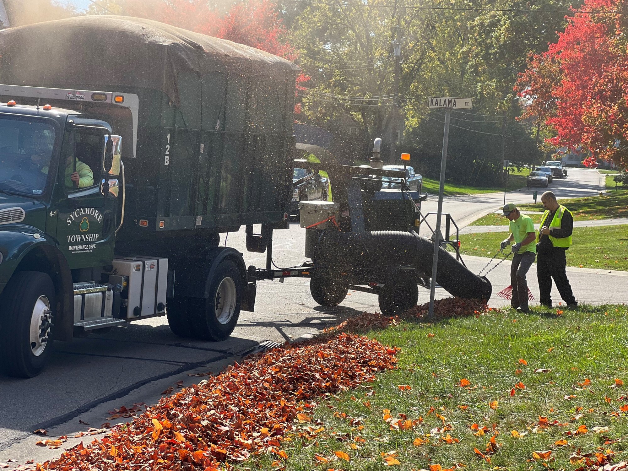 Township maintenance truck parked at a curb while workers collect a large pile of autumn leaves.