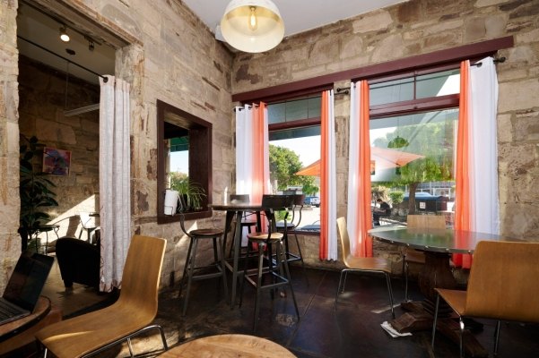 Coffee shop interior with stone walls, high-top tables, and large windows with orange curtains.