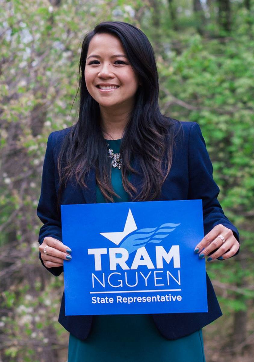 Woman in navy blazer smiling and holding a blue campaign sign outdoors.