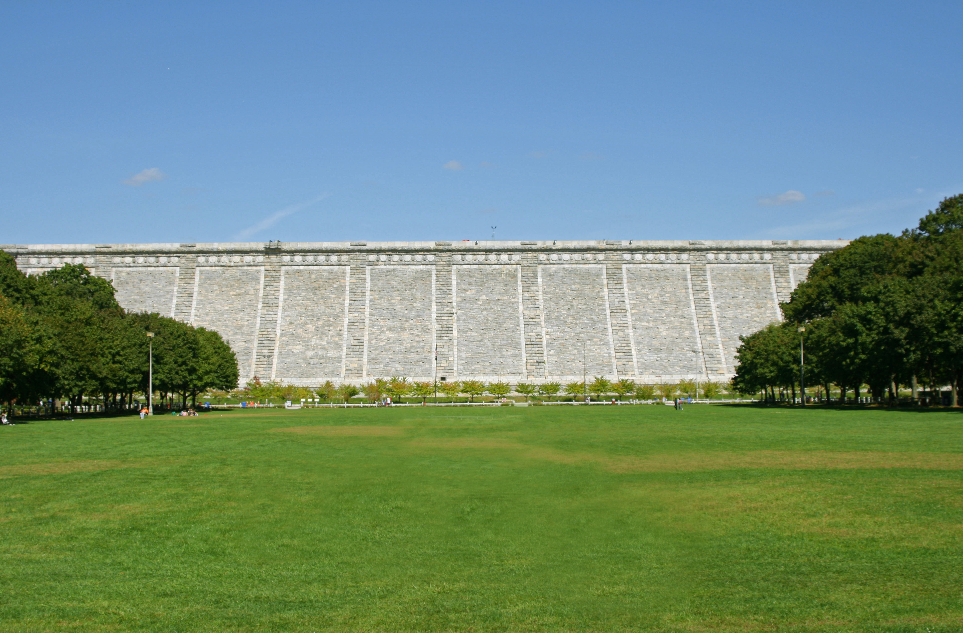 Large stone dam with buttresses viewed from a wide green lawn under a blue sky.
