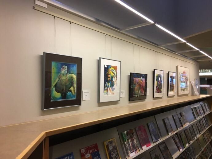 Framed paintings displayed on a white wall above wooden magazine shelving in a library gallery.