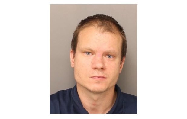 Mugshot of a man with short brown hair wearing a dark blue shirt against a gray background.