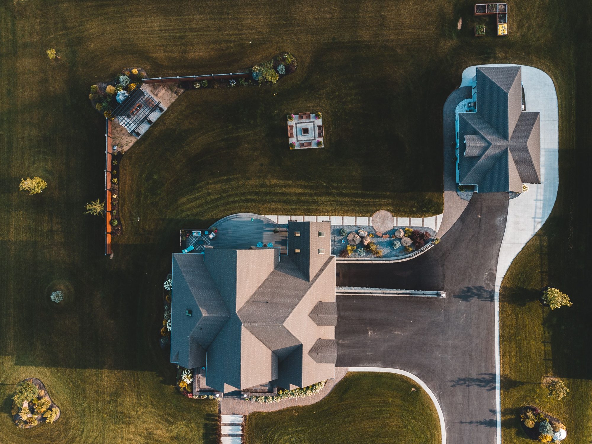 Aerial drone view of a large suburban home with an extensive paved driveway, patio and pool deck surrounding a comparatively small area of lawn.