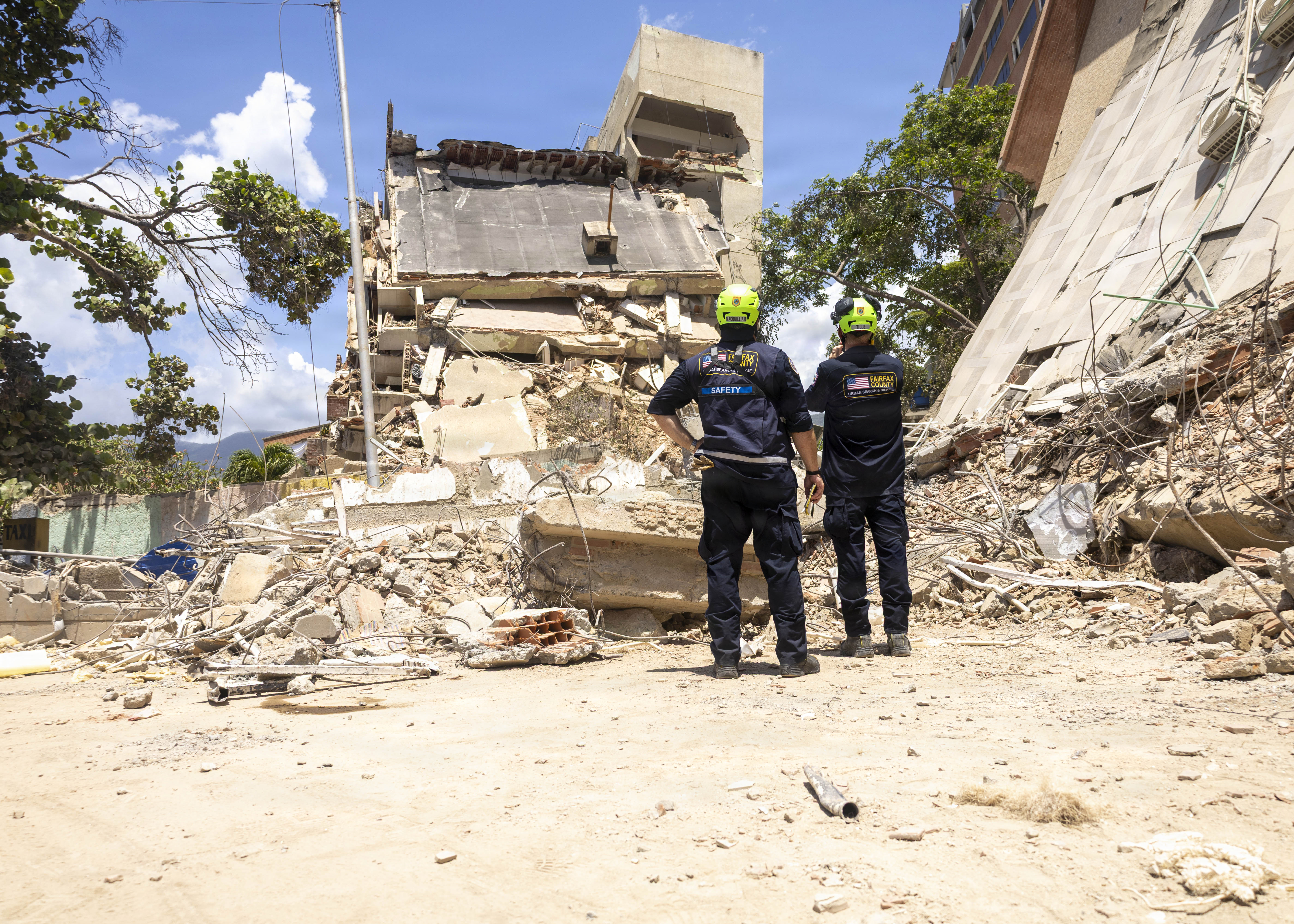 Two rescue workers in helmets standing before a collapsed multi-story building surrounded by rubble.