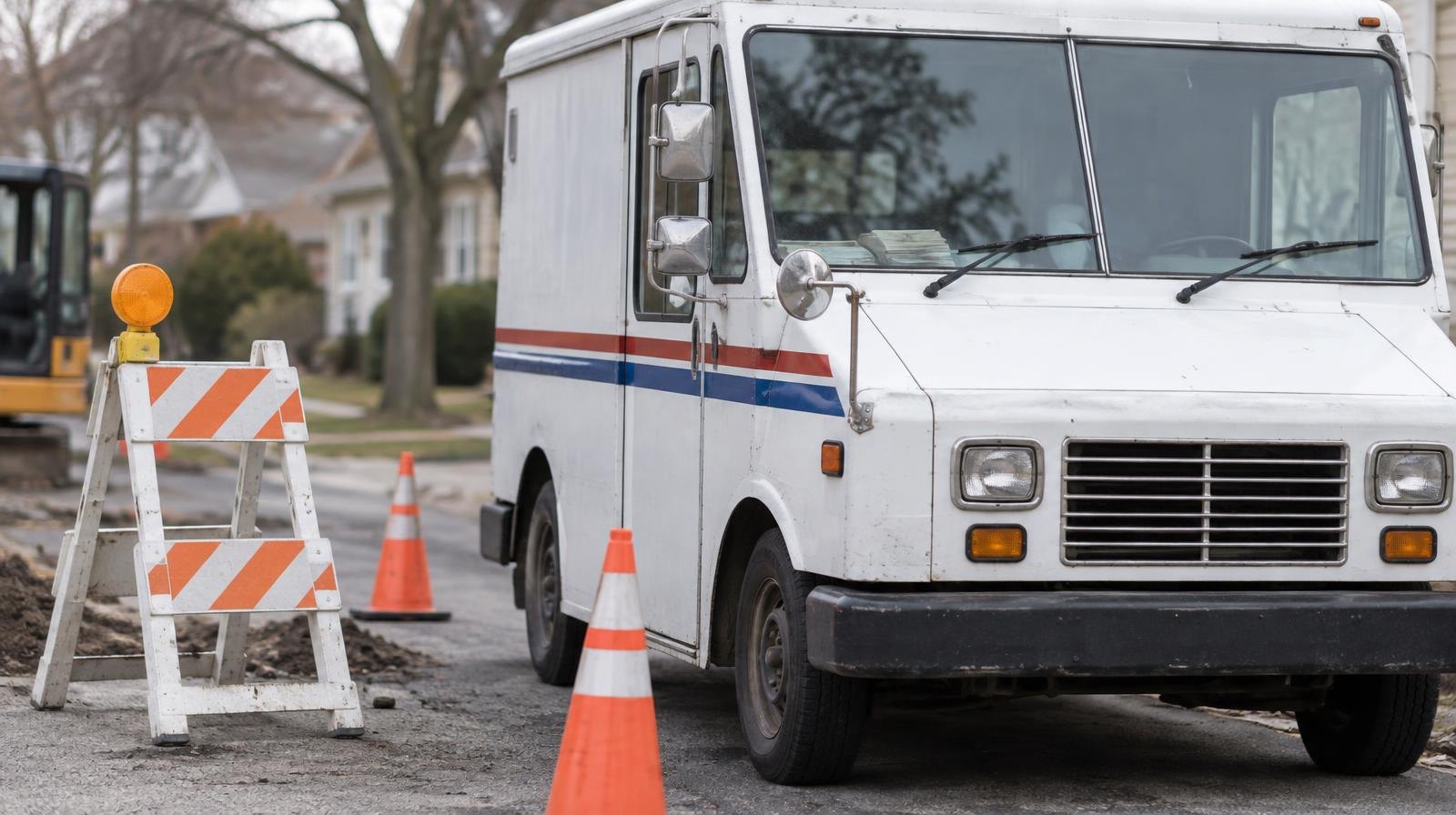 White postal delivery truck parked next to orange traffic cones and a road construction barricade.