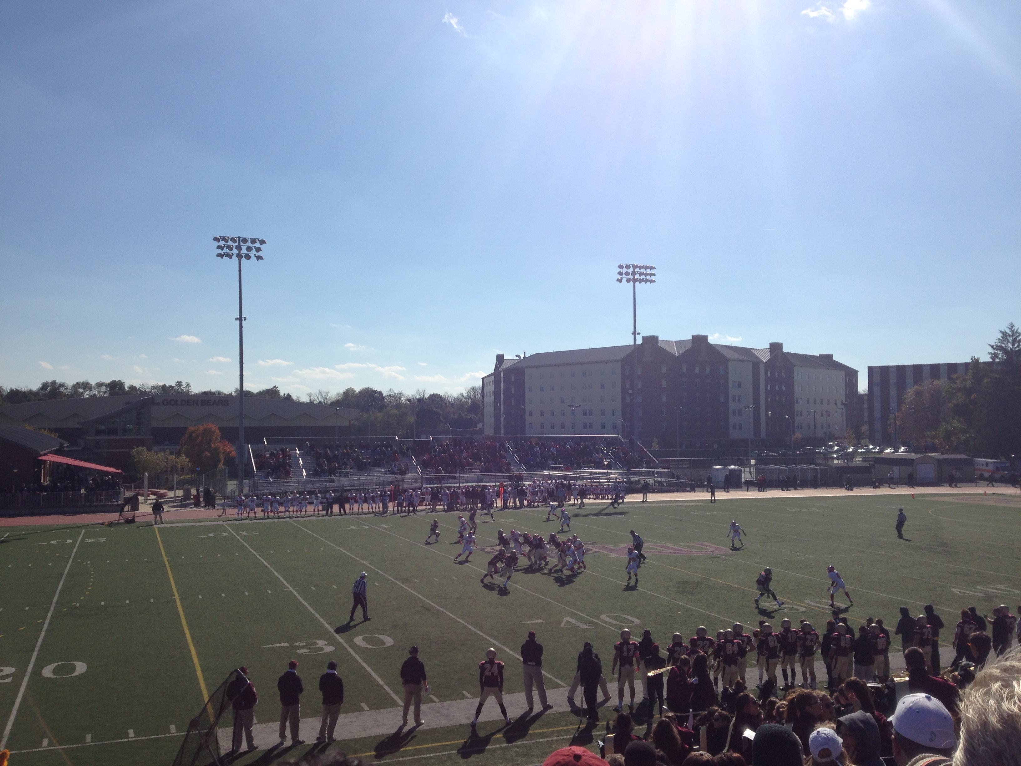College football game on a turf field with players and sideline spectators under stadium lights.