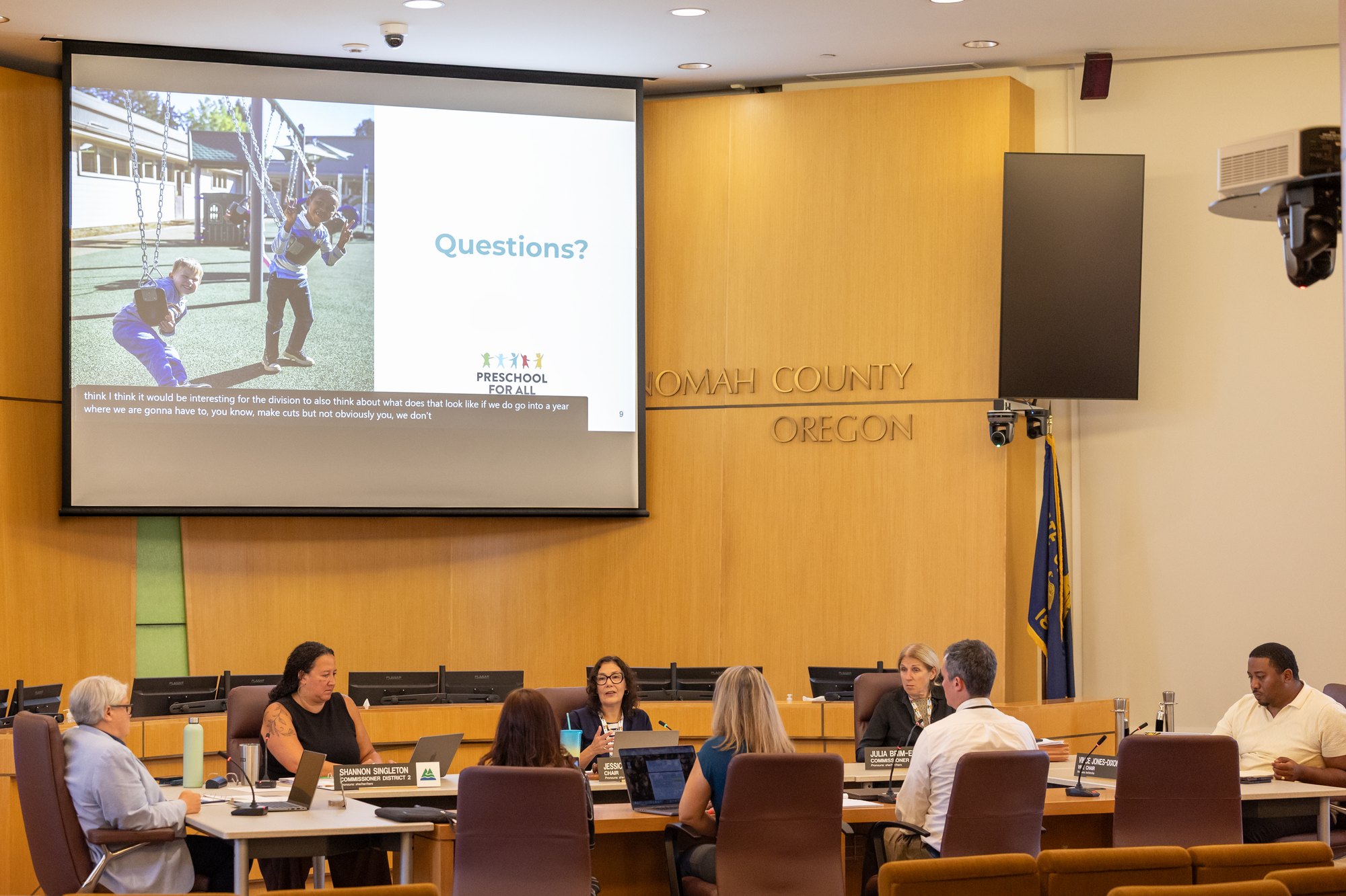 People seated at a curved table in a government board room with a Preschool for All slide projected on screen.