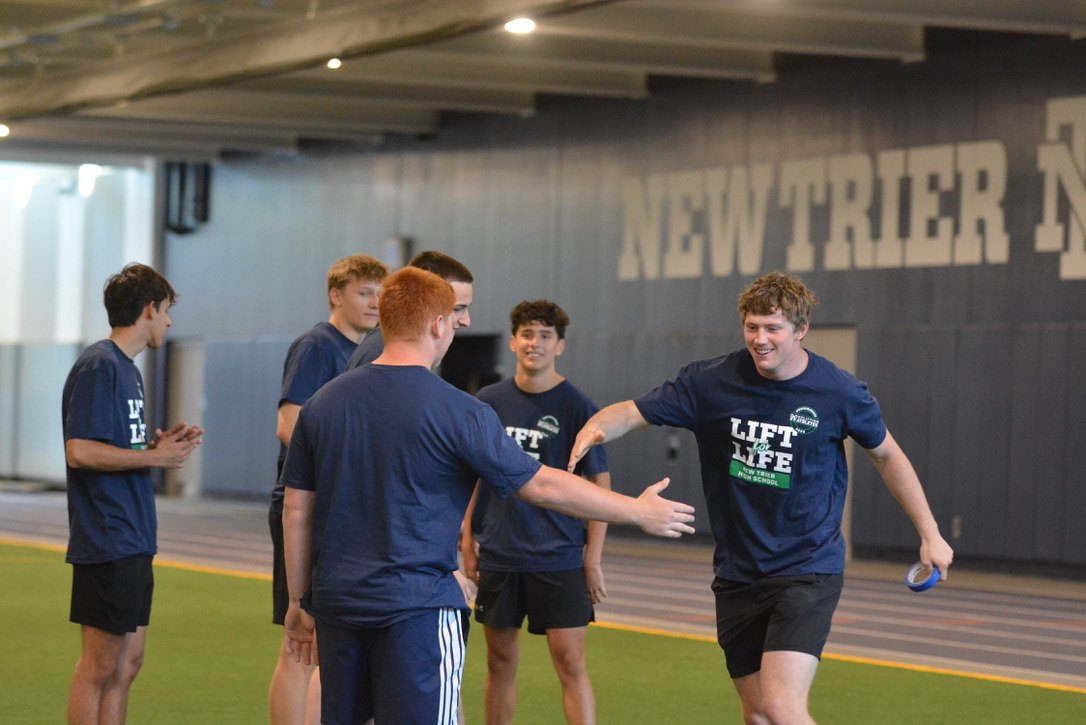 Young athletes in dark Lift Life t-shirts greeting each other in an indoor fieldhouse with green turf.