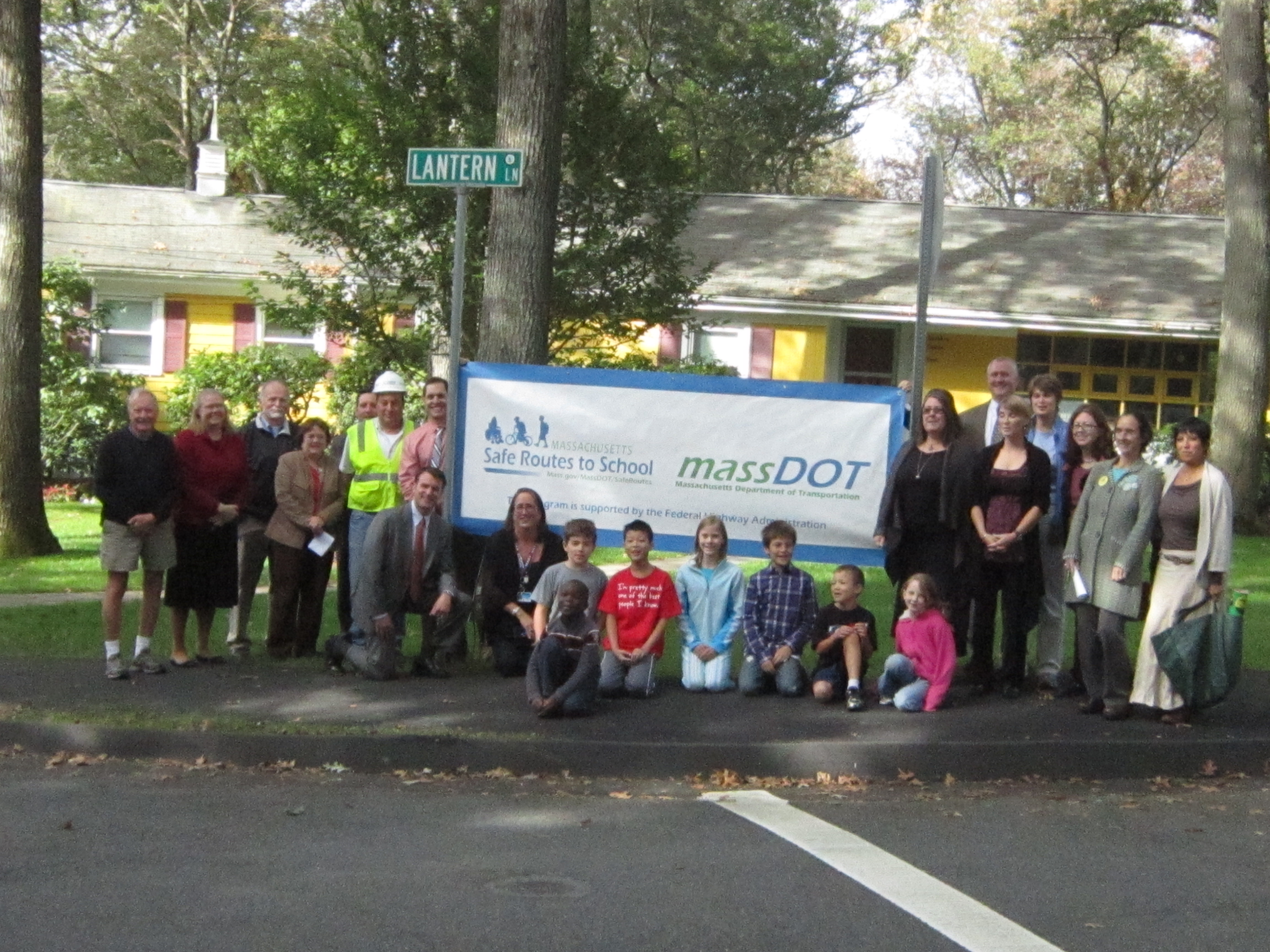 Adults and children posing outdoors in front of a Safe Routes to School banner near a school.