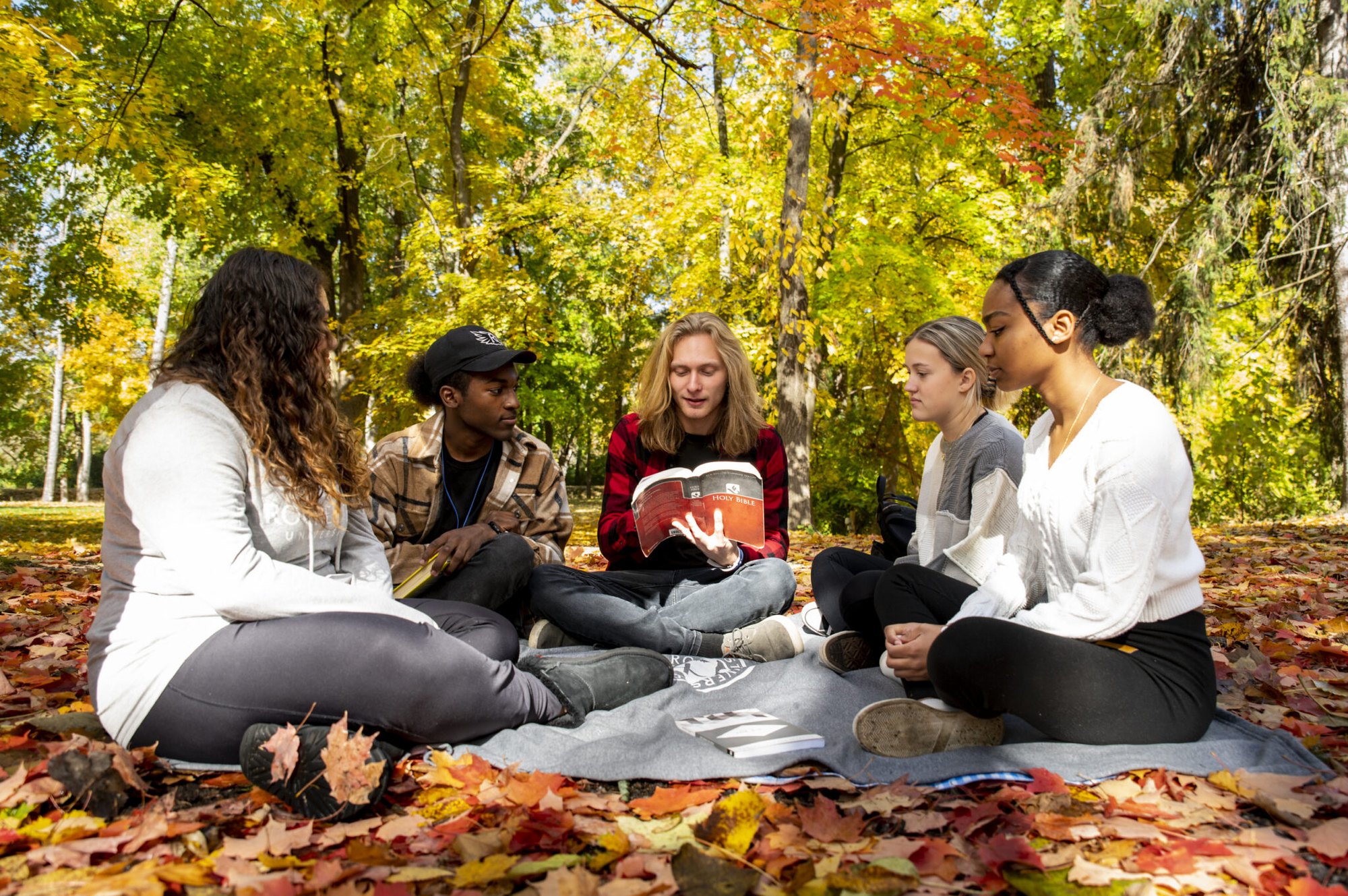 Five students sitting in a circle on a blanket among autumn leaves, one holding an open book.