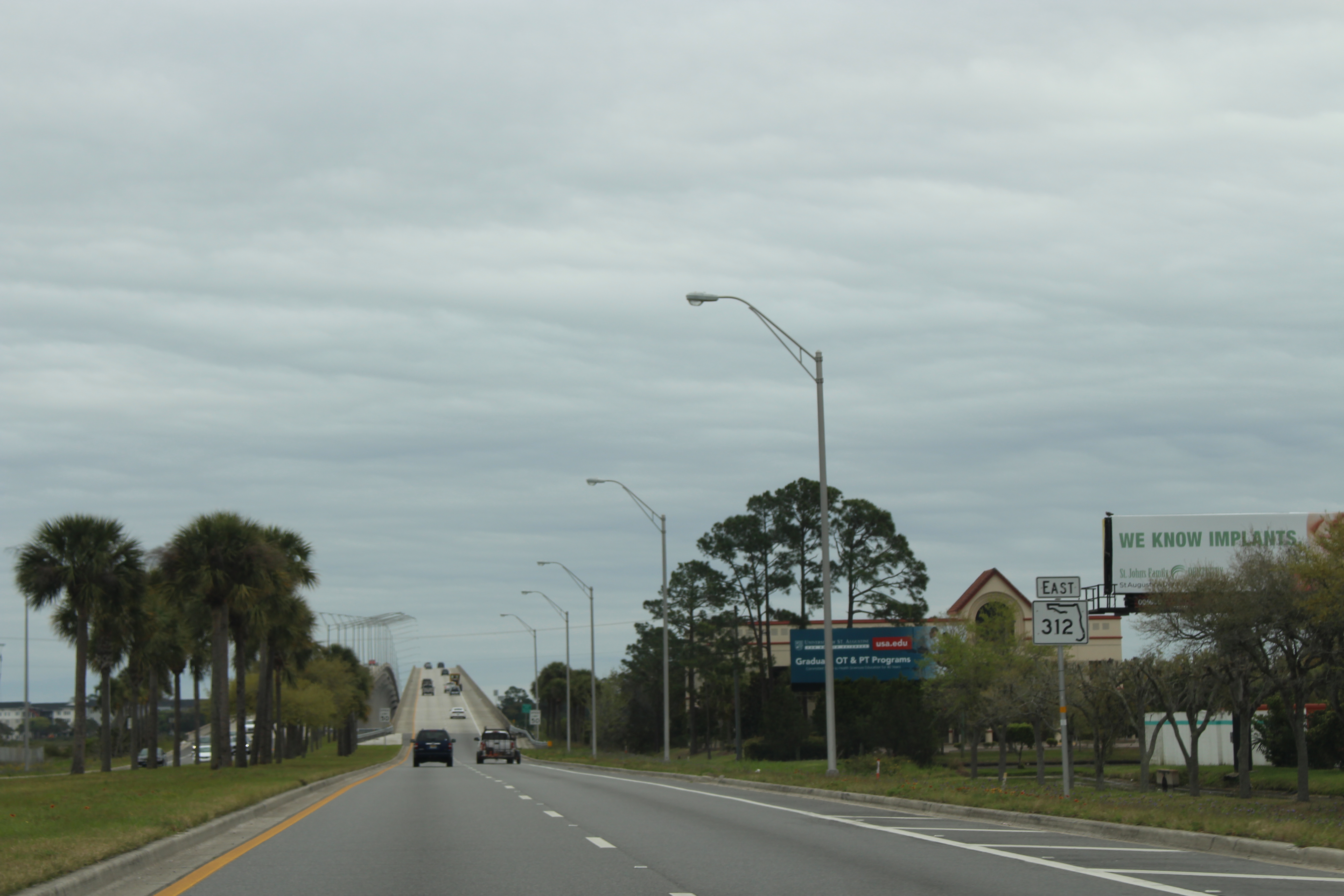 A multi-lane road lined with palm trees leading toward a bridge under overcast skies.