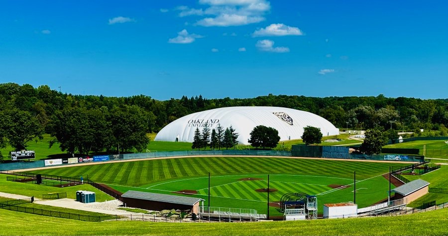Aerial view of a striped grass baseball diamond with bleachers and a white dome structure behind the outfield.