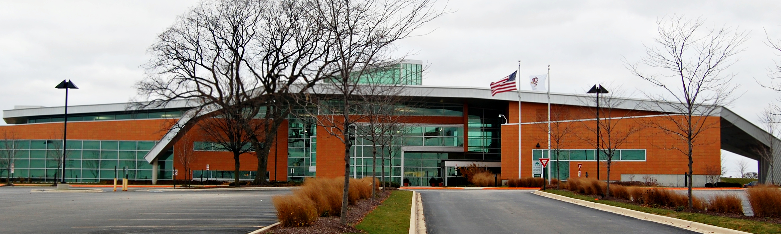 Modern brick and glass library building with two flagpoles and a parking lot in front.