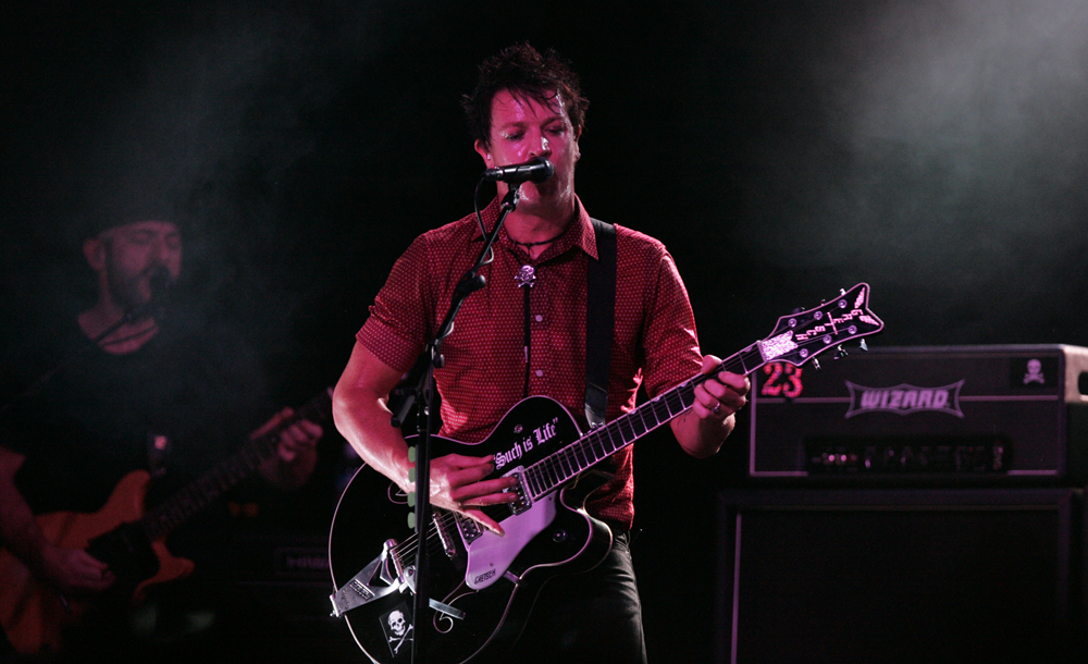 Guitarist sings into a microphone on a stage lit with pink and purple lights.