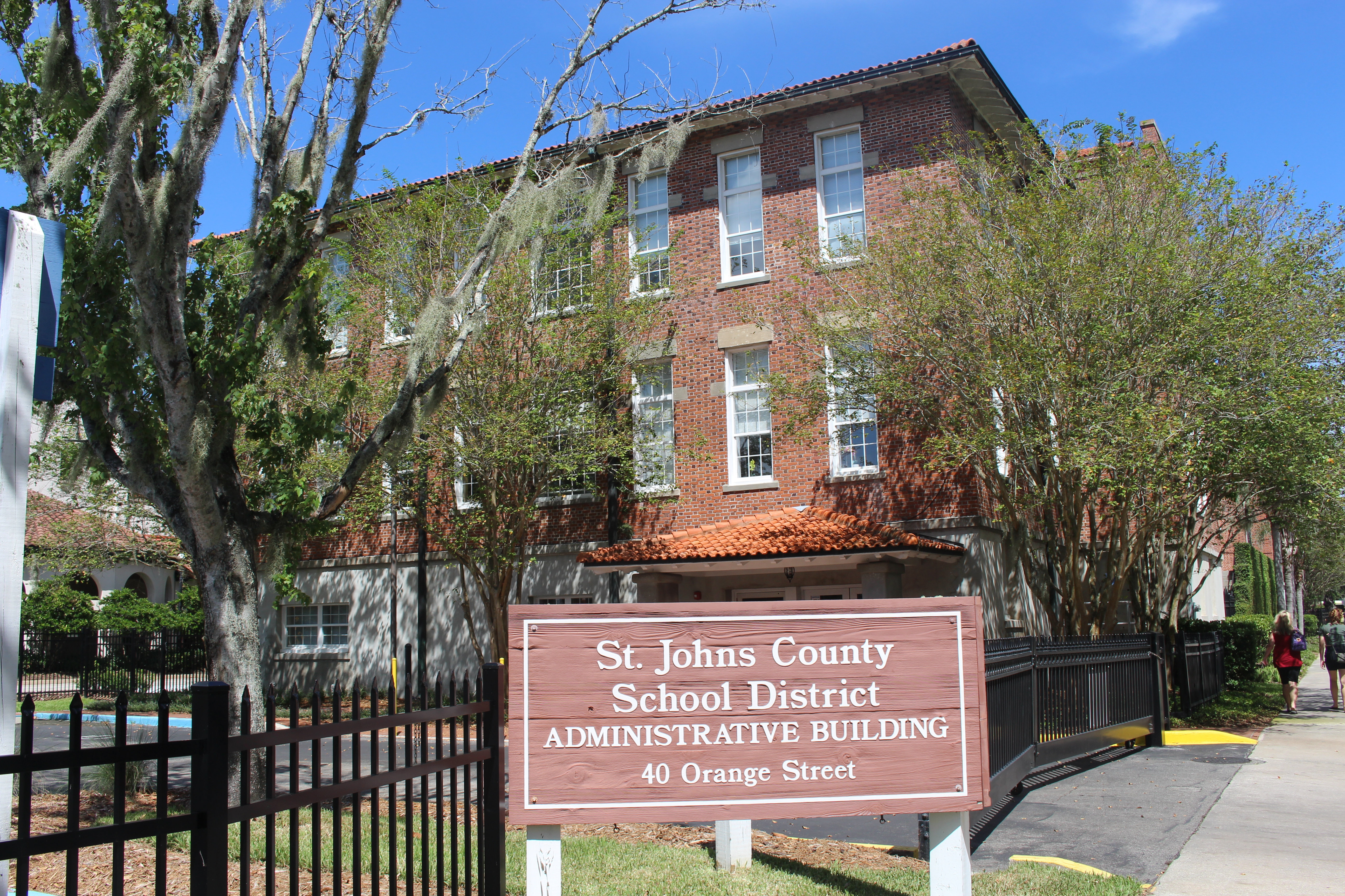 Red brick school district administrative building behind a black iron fence, with Spanish moss trees.