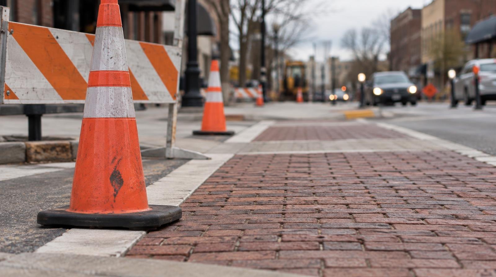Brick paver crosswalk at an intersection with orange construction cones and a barrier at the edge.