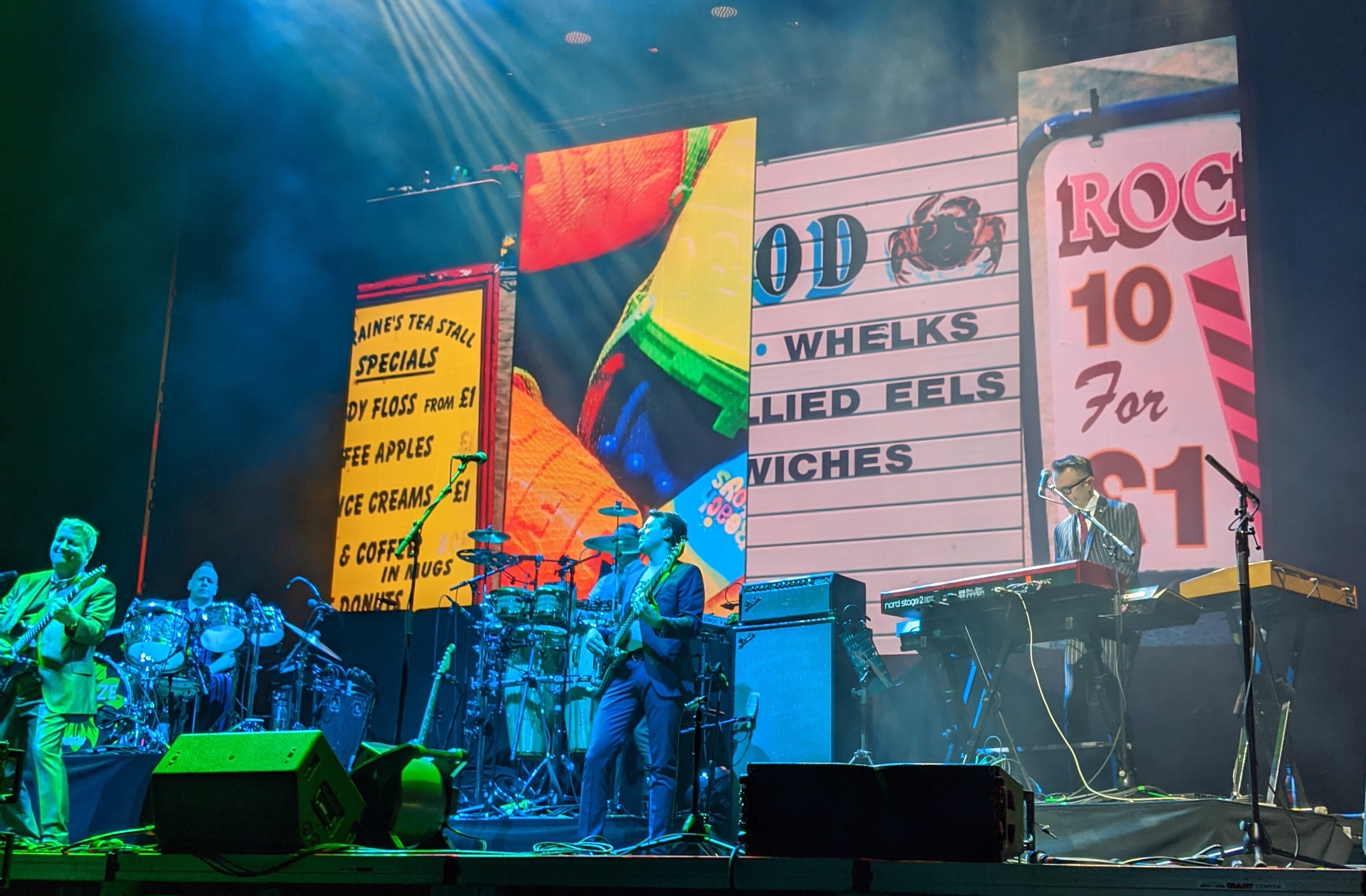 Band members performing on a lit stage with large colorful backdrop screens.